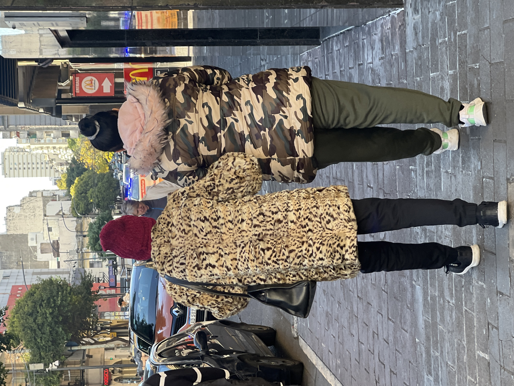 Two people stand on a brick sidewalk next to a busy street in Buenos Aires, Argentina. The first person wears a camo jacket with a pink-trimmed hood, dark green pants, and white and green sneakers. The second person wears a leopard-print coat, black pants, black sneakers with white soles, and a red knit hat. They both appear to be looking at something in front of them on the street. Cars are parked along the street, and traffic passes. Buildings line the street, with trees visible in the background. A McDonald's sign is visible in the top left of the frame.