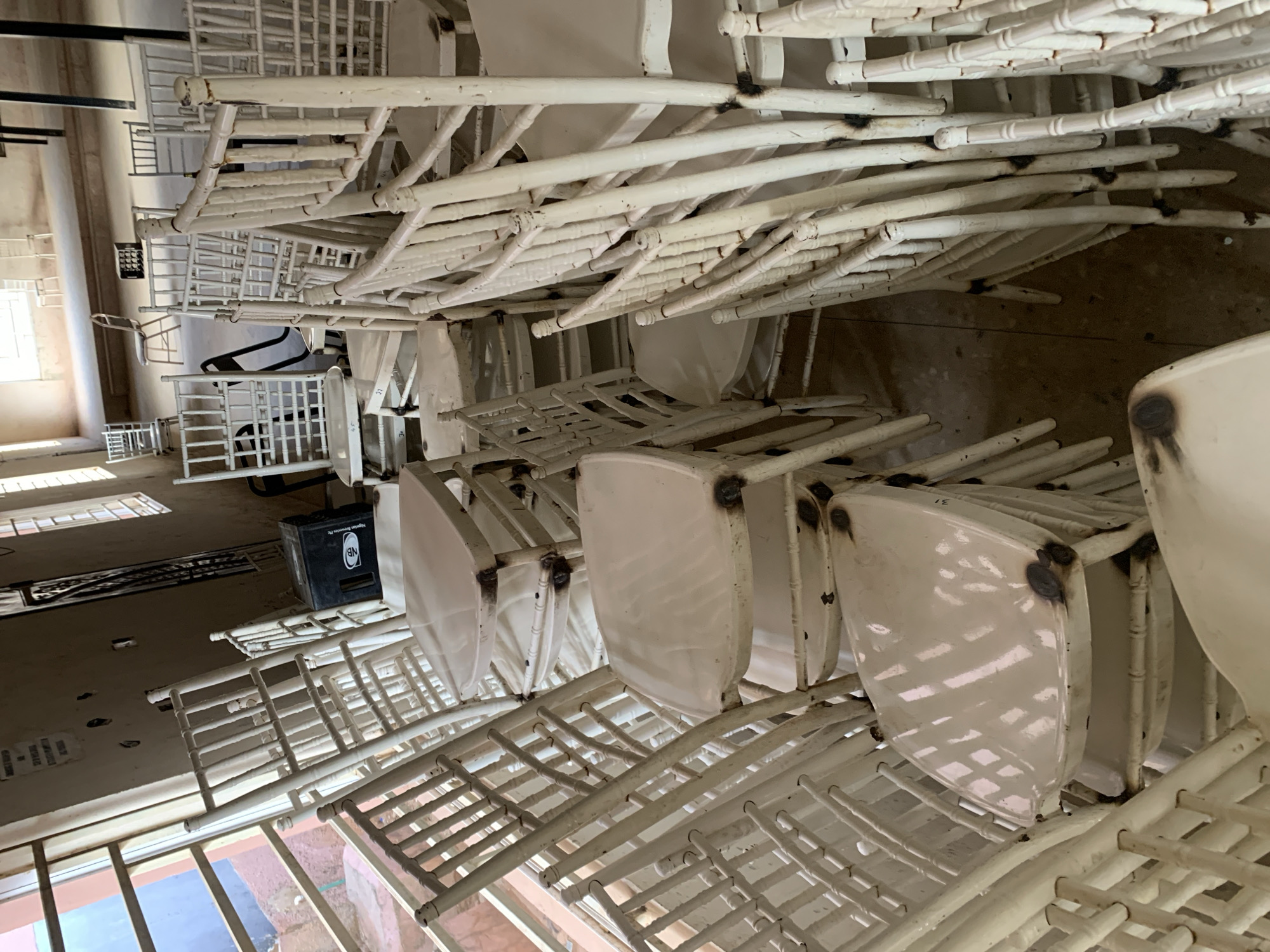 The image shows a room in a building filled with stacks of white chairs. The chairs appear to be made of metal and plastic. They are stacked on top of each other in a disorganized manner. A black box with the letter "NB" is visible in the background of one of the areas between stacked chairs. Based on the text, the location is the New Stadium in Nigeria.