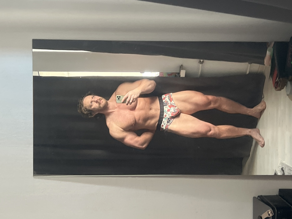 A shirtless man with curly hair and a mustache poses for a mirror selfie. He wears patterned underwear and a phone in his left hand. His body is muscular, with defined biceps, pectoral muscles, and legs. He stands against a dark background, likely a curtain. Part of a white wall is visible on the left and the lower right shows a desk or table, and some items on the floor.