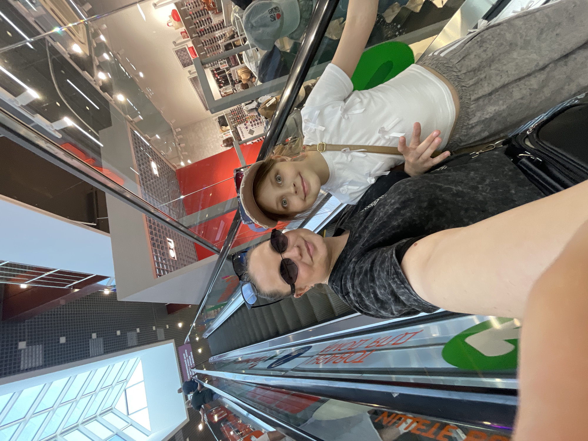 A selfie captures two individuals on an escalator, presumably in the city of Izhevsk, Russia. The foreground is dominated by the arm of the person holding the camera. The escalator's metallic surface with direction arrows is visible. The two people in the image, presumably a mother and daughter, are ascending. The daughter, positioned behind the adult, is wearing a white blouse with bows, grey pants, and a light brown cap. The woman taking the photo has dark sunglasses, a dark-colored shirt, and another pair of sunglasses propped on her head. The background reveals the architecture of a shopping mall, with shops, displays, and a glass ceiling.