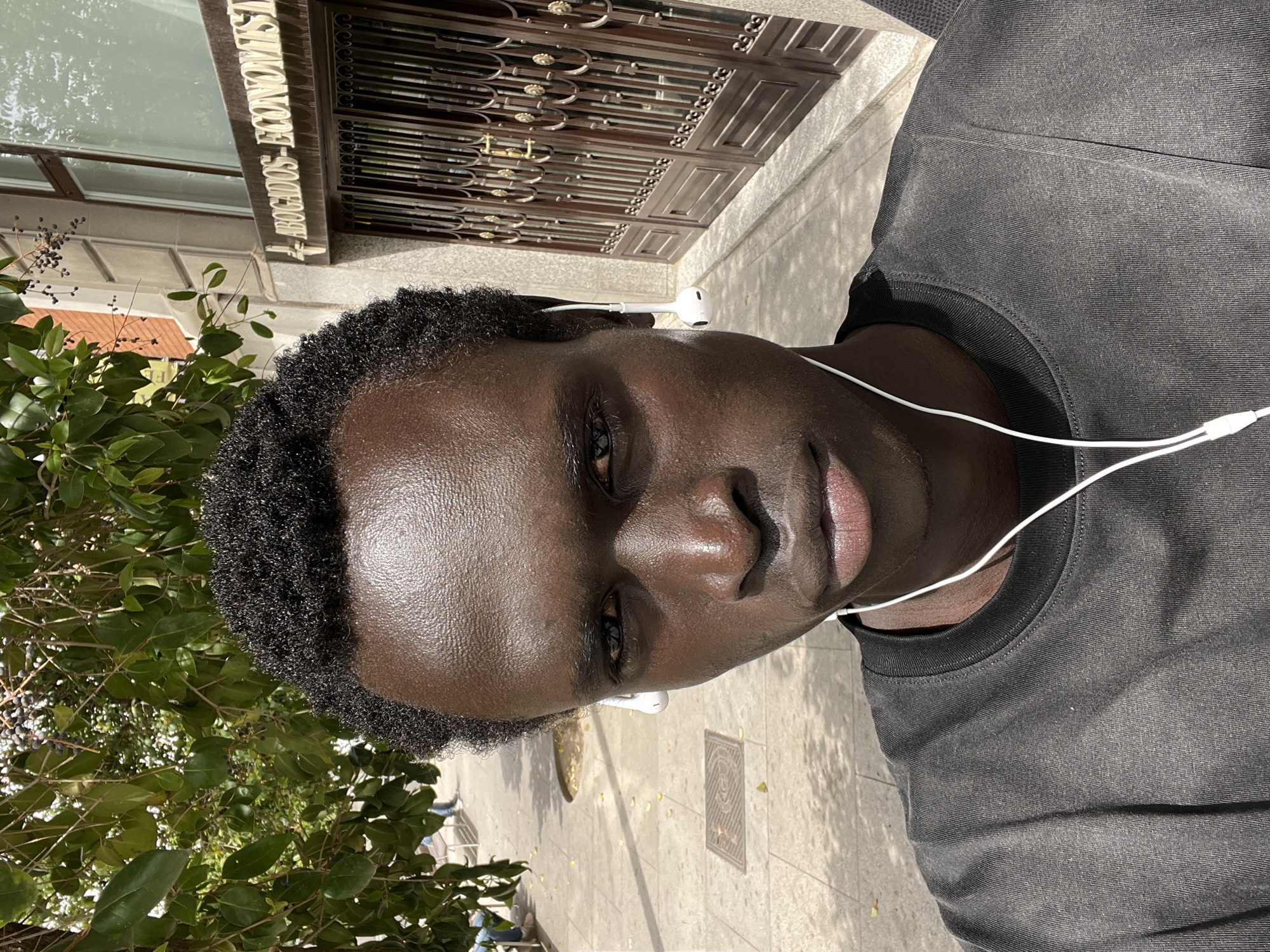 A close-up, high-angle selfie captured outdoors on a sunny day. The image is rotated 90 degrees counter-clockwise. The primary subject is a person with dark skin and short, curly black hair, looking directly at the camera with a neutral expression. They are wearing a dark grey t-shirt and white wired earbuds, one visible in each ear.
The background reveals an urban setting. To the left, there's lush green foliage from a tree or bush, featuring dark berries. Behind the person, a building facade is visible with a large glass window reflecting the surroundings. Below the window, a sign bearing the white text "ABOGADOS - ECONOMISTAS" (Spanish for "Lawyers - Economists") is prominently displayed. Adjacent to this, an elaborate, dark brown metal gate or door with intricate scrollwork and gold accents is present. The ground beneath is a light-colored concrete sidewalk, showing strong shadows indicative of bright sunlight and a visible square metal grate. The overall setting is consistent with a street in Madrid, Spain.