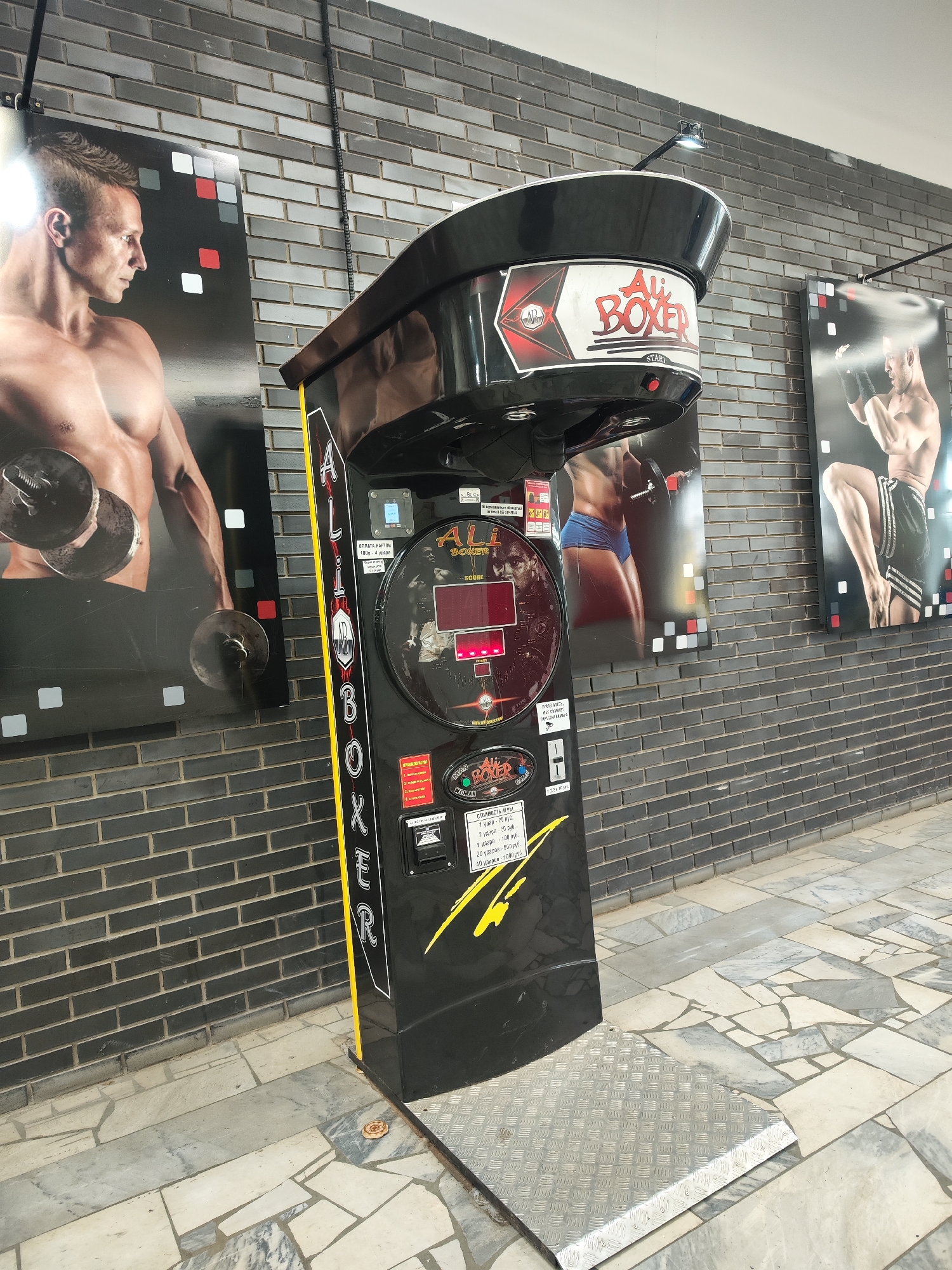 The image shows an "Ali Boxer" punching arcade machine in what appears to be a building in Chaikovsky, Russia. The machine is black with yellow accents and features the "Ali Boxer" logo prominently. The machine also displays pricing information for different numbers of hits in Russian rubles. It's placed on a silver metallic platform, and the floor is paved with irregular light-colored tiles. Behind the machine, the wall is clad in dark gray bricks and is decorated with three posters of muscular individuals: one holding weights, one posing, and the third appearing to box. A black pipe runs along the wall near the ceiling.
