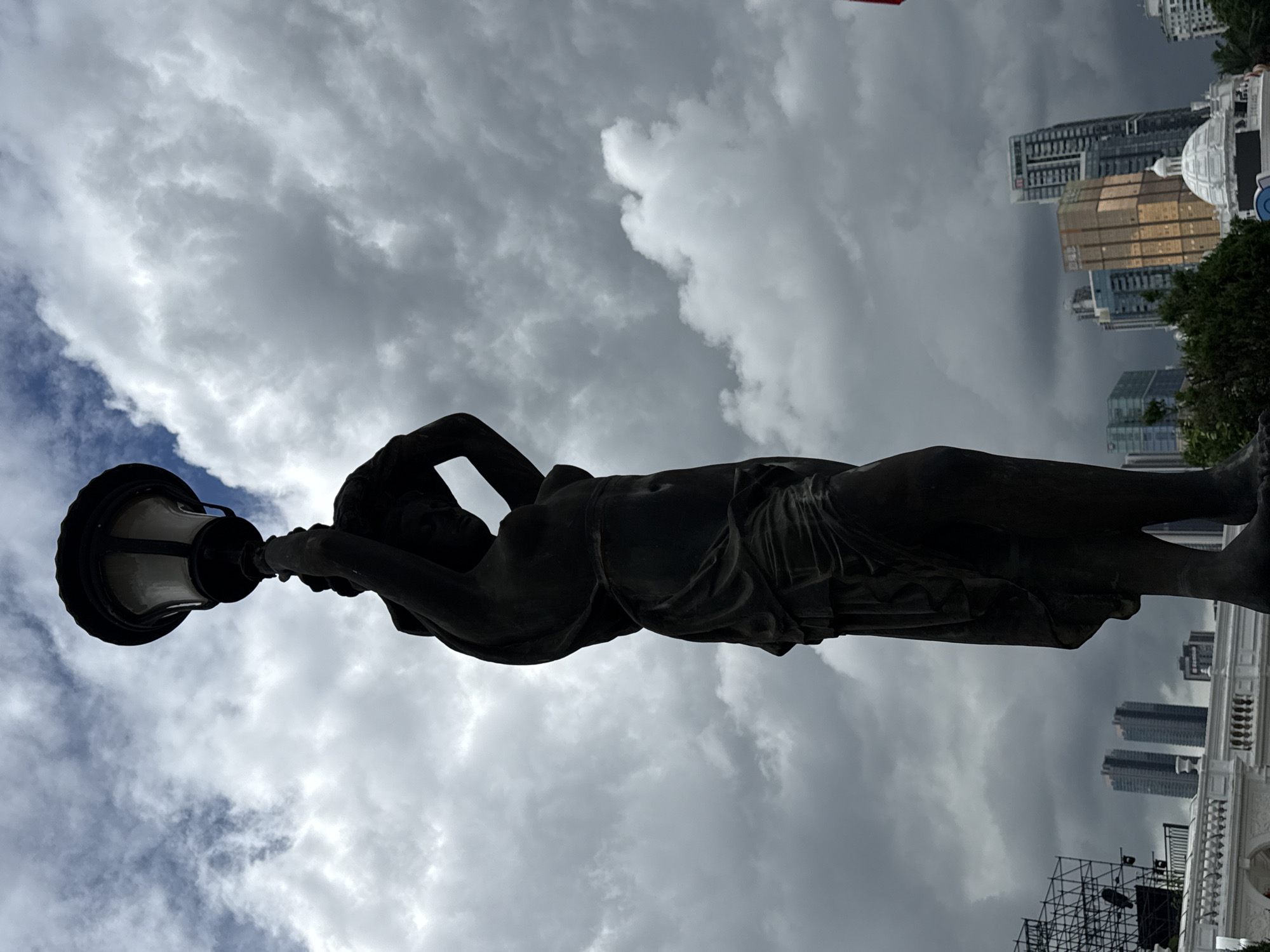 A low-angle, full-body shot captures a dark bronze statue of a draped female figure holding a lantern aloft against a dramatic cloudy sky. The statue is positioned in the foreground, with its silhouette dominating the frame. Behind the statue, the cityscape of Nanshan District, China, is visible. This includes several modern high-rise buildings, some with reflective facades, as well as a white domed structure. To the far left, scaffolding from a construction or event setup is visible. The lighting suggests it is daytime, but the overcast sky creates a somewhat moody atmosphere. The statue appears to be a decorative element in an outdoor public space.