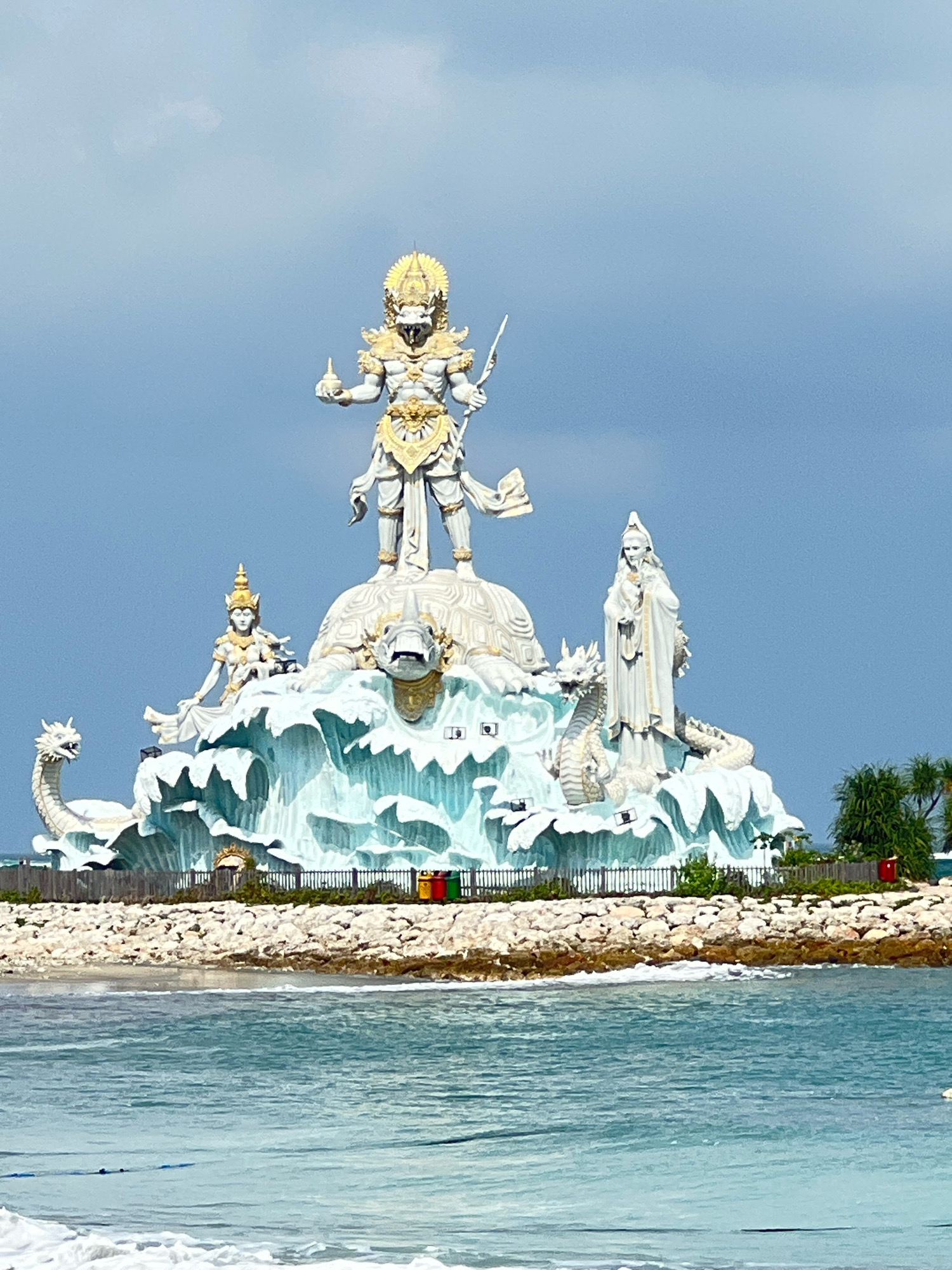 The image shows the Garuda Wisnu Kencana Cultural Park in the Ungasan peninsula, south of Kuta, Indonesia. The centerpiece of the park is a massive statue of the Hindu deity Vishnu riding on the back of Garuda, a mythical bird-like creature. The statue is depicted in a dynamic pose, with Vishnu holding a spear and Garuda with outstretched wings. The sculpture is set against a backdrop of a partly cloudy sky. In front of the main statue, there are smaller sculptures of mythical creatures and celestial beings, including dragons and what appear to be gods or goddesses. The entire structure is built on a tiered platform resembling waves and sea creatures. The base of the monument is surrounded by a rocky shoreline and a body of water, likely the ocean, with a fence and some greenery visible in the background. The image captures the grandeur and intricate details of the sculpture, highlighting its cultural and religious significance.