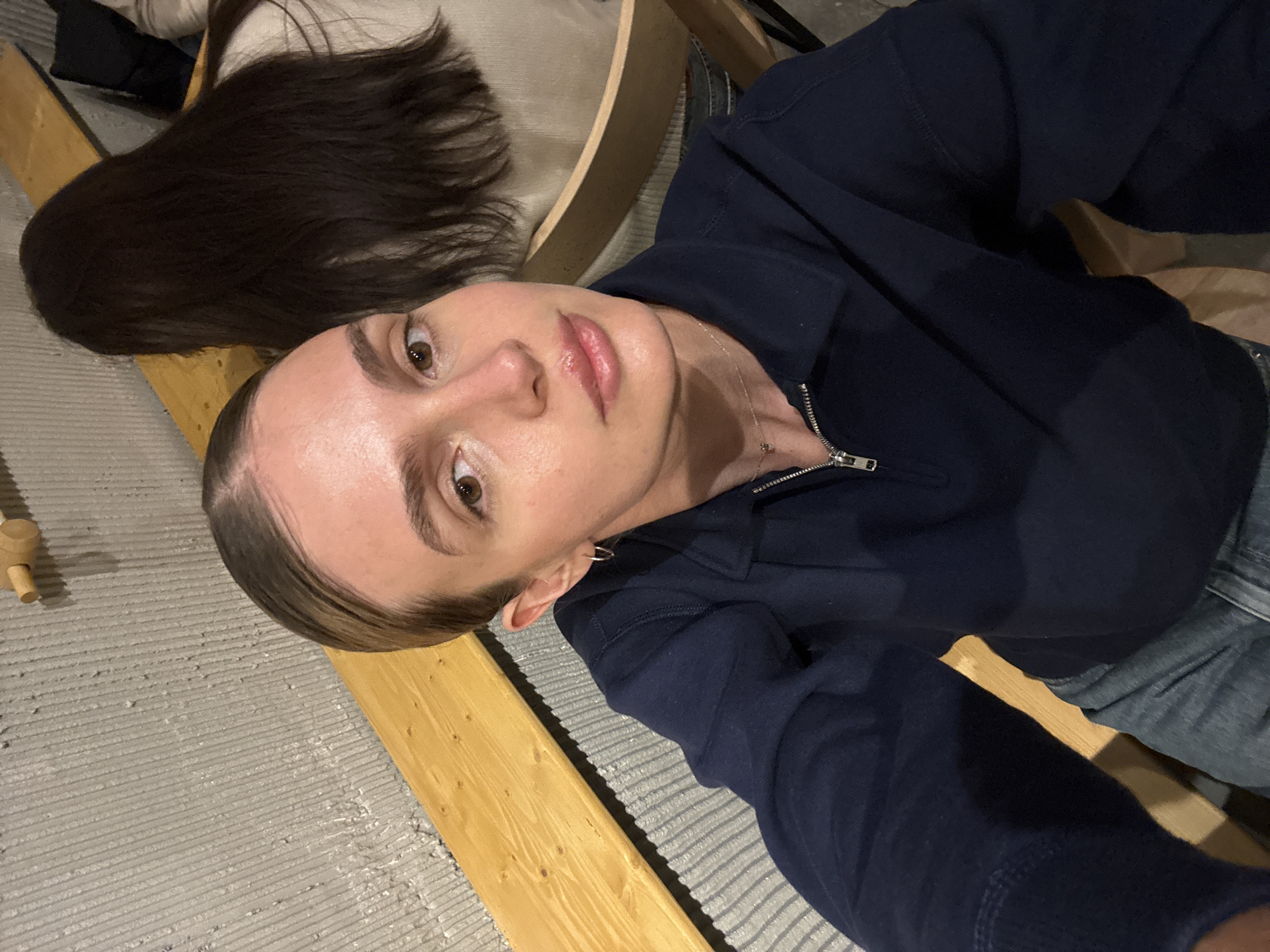 A selfie is taken from a low angle, focusing on the face of a light-skinned person with light brown eyes and slicked-back light brown hair. They are wearing a dark blue zip-up sweatshirt and a thin silver necklace. An earring is visible in their left ear. The background consists of light gray textured flooring, a wooden beam, and brown hair belonging to another person. Some clothing and furniture are also visible in the background. There is no information to specify that the image was taken in Tbilisi, Georgia.