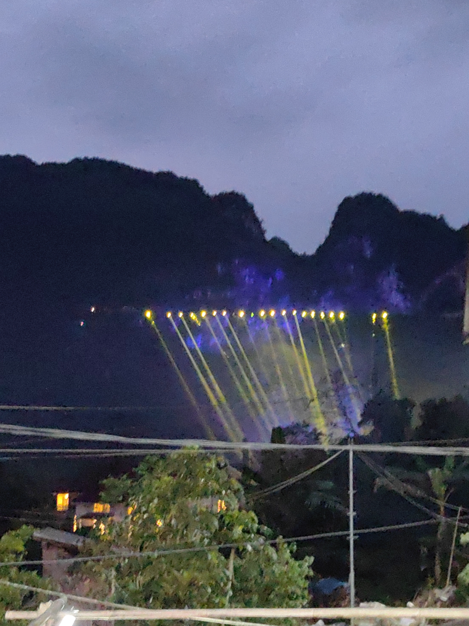 The image shows a nighttime view in Xã Đàm Thủy, Vietnam. The foreground features trees and utility poles with wires. In the midground, a building with lit windows is partially visible through the trees. The background consists of dark, hilly terrain illuminated by multiple spotlights. The lights are arranged in a line and project yellow beams onto the rock formations. Additionally, there is some blue light also illuminating the hills. The sky is cloudy and dark.