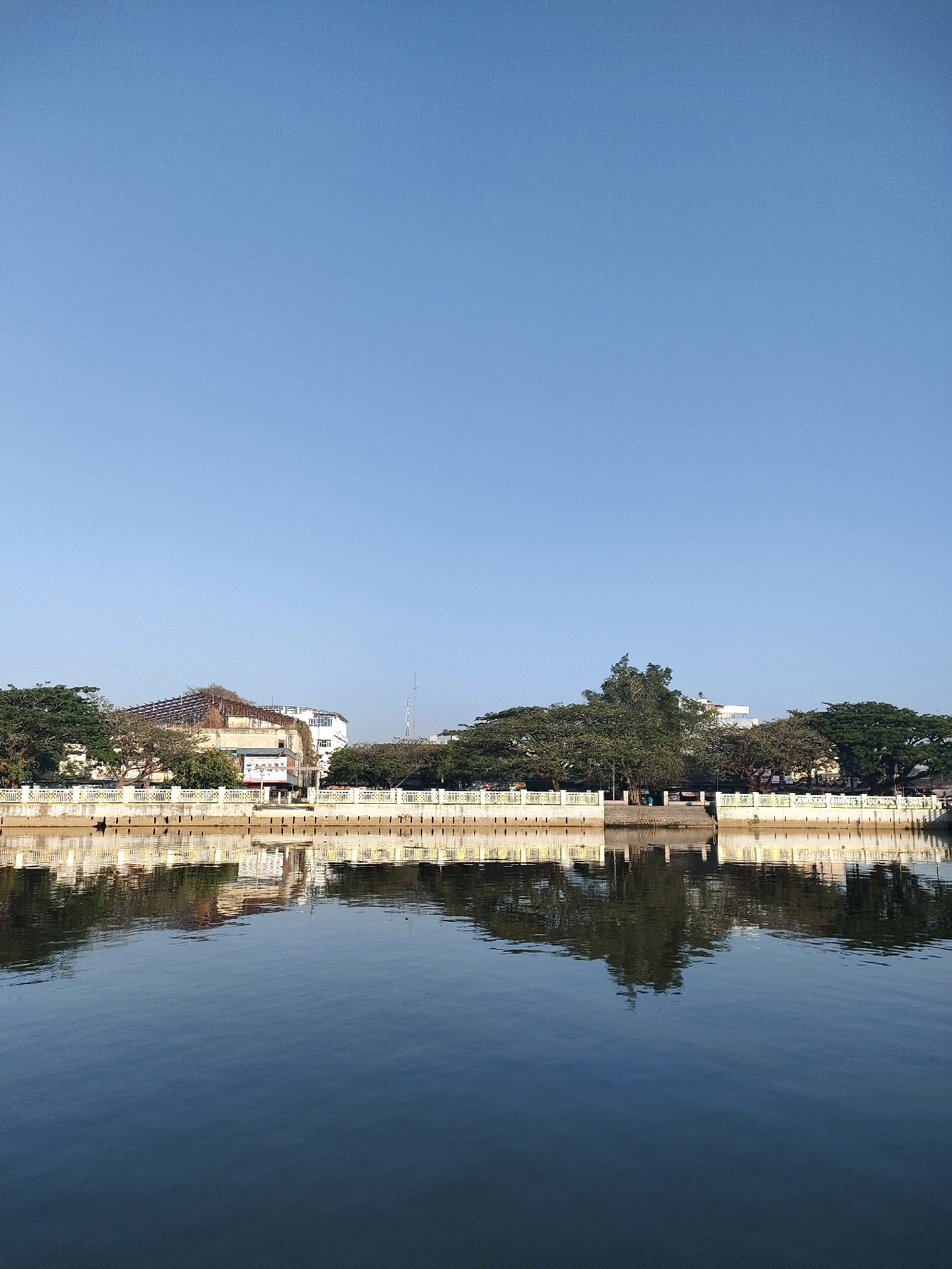 A scenic view of the Martapura River in Banjarmasin, Indonesia, showcases the city's waterfront. The clear blue sky dominates the upper portion of the image, creating a serene backdrop. Below, a white embankment with a yellow railing lines the river, separating the water from the buildings and trees on the shore. The buildings are a mix of old and new, with some appearing to be under renovation. Lush green trees provide shade and add to the picturesque setting. The river itself is calm and reflects the structures and vegetation on its surface, creating a mirror-like effect. The overall scene evokes a sense of tranquility and highlights the natural beauty of Banjarmasin's urban landscape.