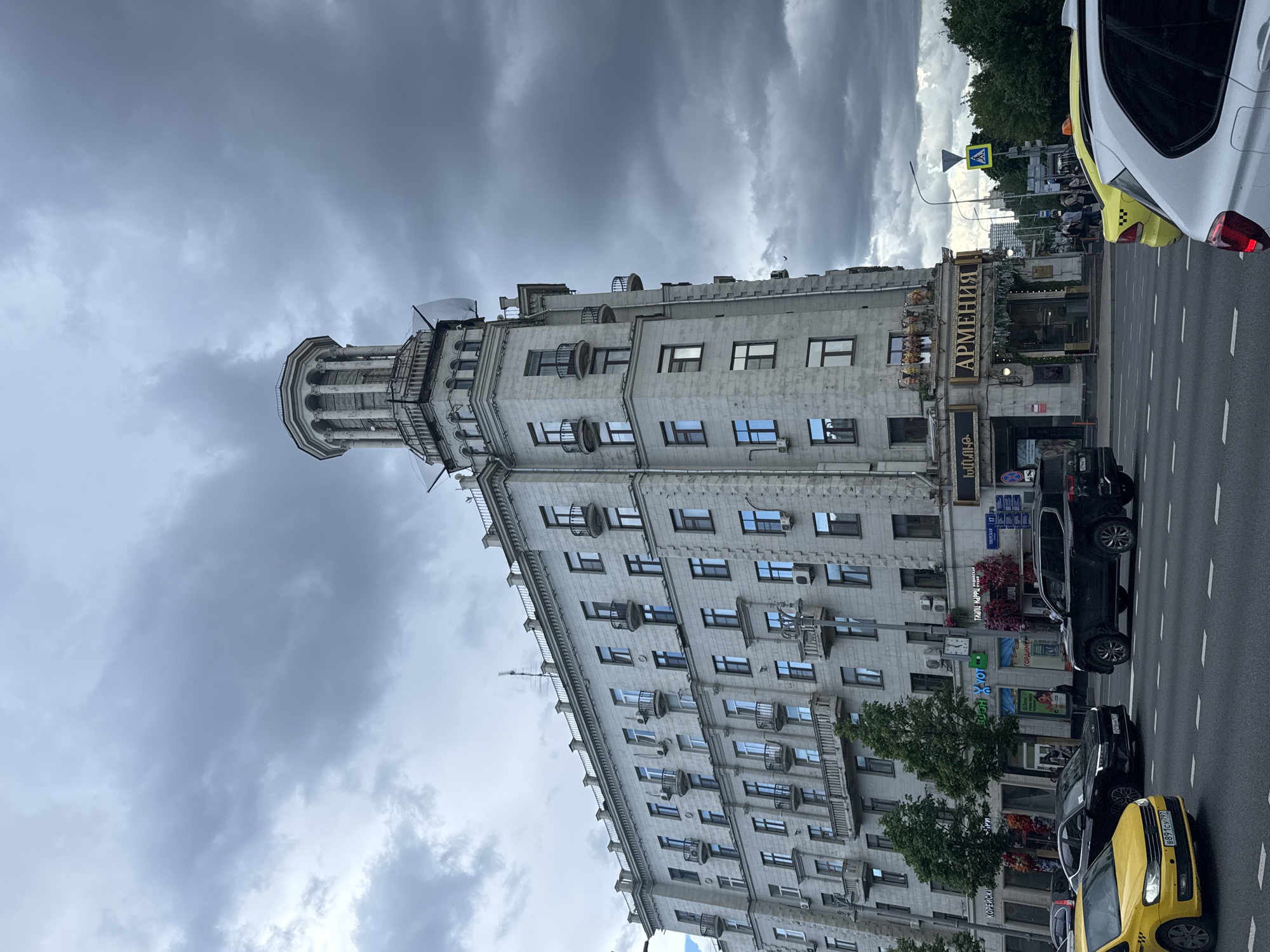 The image shows a building with a distinctive tower in Moscow, Russia. The building has multiple floors and a light-colored facade with numerous windows and small balconies. The restaurant "Armenia" is visible at the base of the building. Several cars are parked or driving on the street in front of the building, including a yellow taxi. The sky is overcast with dark, heavy clouds. Traffic signals and street signs are also visible in the background.