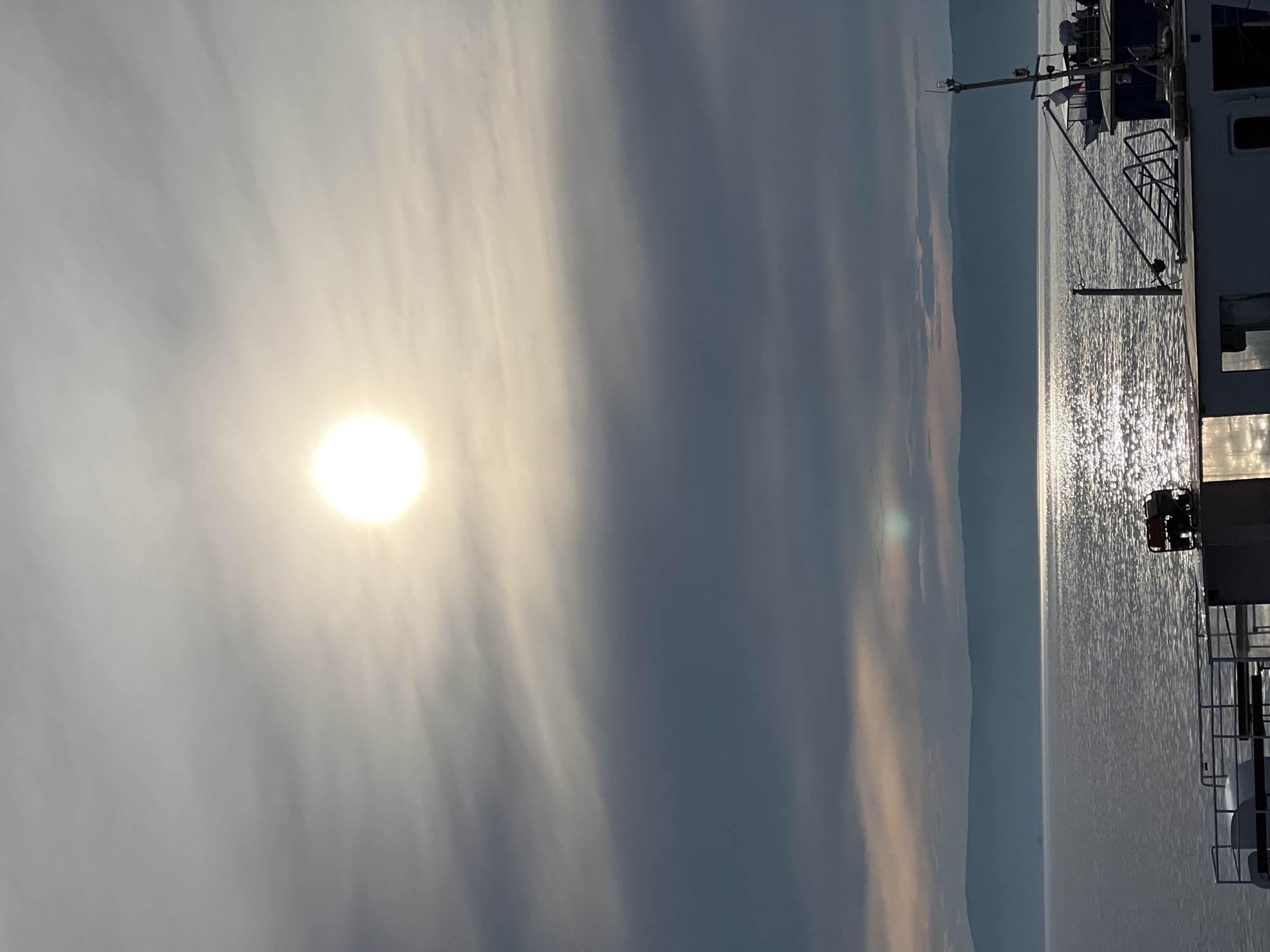 The image depicts a wide, calm body of water, likely Lake Baikal, with a distant, hazy mountain range forming the horizon. The sky is overcast with layers of clouds, but a bright, circular sun is visible near the top center, casting a luminous glow that reflects on the water's surface. In the foreground, the edge of a boat is visible, featuring white railings and a portion of a cabin. A piece of machinery, possibly a generator, sits on the deck of the boat. Another, smaller boat is docked to the right, its mast and a small flag are visible. The scene suggests a serene, possibly early morning or late afternoon setting in Listvyanka, Russia, a popular tourist village on the western shore of Lake Baikal. There are no people clearly visible in the image.