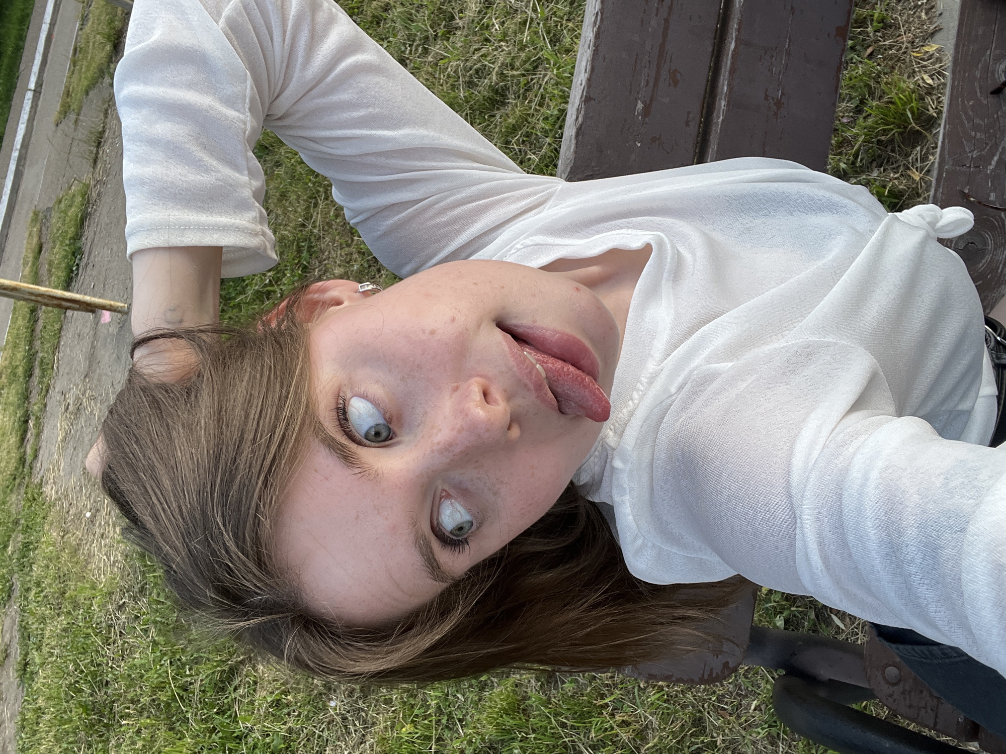 This is a medium shot, close-up, outdoor, low-angle, horizontal photograph of a young woman lying on her back on a wooden bench. She is wearing a sheer, long-sleeved white shirt. Her head is tilted back and to the left, with her eyes wide open and looking upwards, and her tongue sticking out. Her hair, which is brown and shoulder-length, is spread out around her head. She has freckles across her nose and cheeks. The bench appears to be in a park or outdoor recreational area, with grass visible beneath and around it. The lighting suggests it is daytime, likely sunny. There are no visible text elements in the image. The overall mood is playful and mischievous.