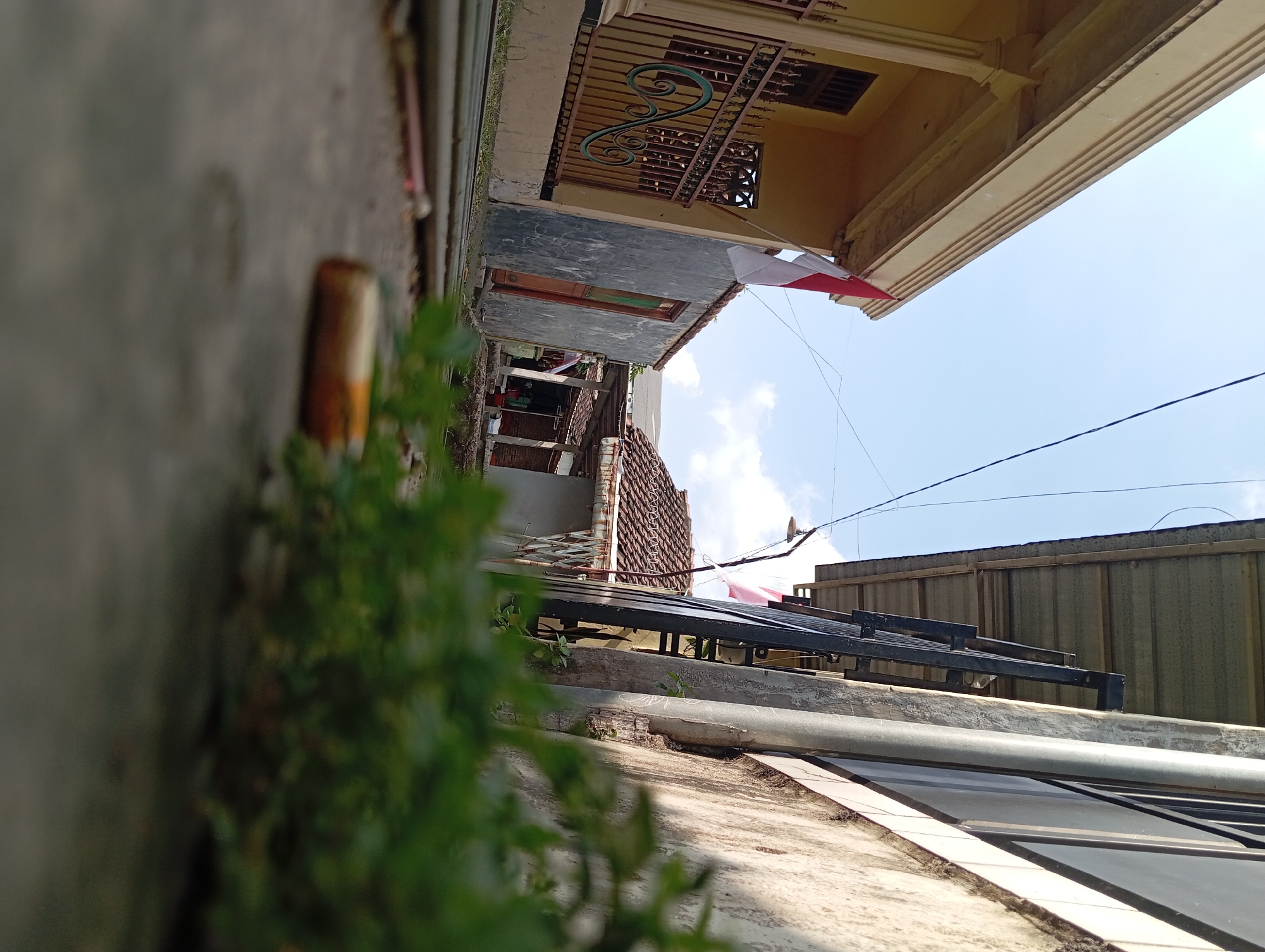 This is a photograph taken from a low angle, looking down a narrow alleyway. On the left side of the alley is a yellow building with a dark-colored fence. The building is decorated with columns and has a window visible. An Indonesian flag is flying from a pole attached to the side of this building.
To the right of the alley, there is a gray building with a simple wooden door. Further down the alley, more buildings with tiled roofs are visible, suggesting a residential area. The ground in the alley is paved with concrete. In the foreground, out of focus, is some green foliage and a cigarette butt lying on the ground. The sky above is blue with some scattered white clouds. The overall scene depicts a simple, possibly residential, street in a town or village.