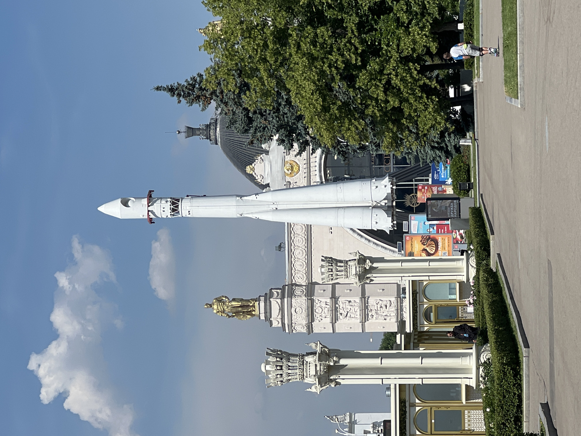 The image depicts an outdoor scene at the All-Russian Exhibition Center (VDNKh) in Moscow, Russia. A large white rocket stands vertically in the foreground, flanked by a golden statue of a man on a pedestal to its left and lush green trees to its right. In the background, a building with a dark dome and architectural embellishments is visible. The sky is blue with scattered white clouds. A person is walking through a manicured garden area, while another person on rollerblades is present in the wider paved area. The overall setting appears to be a sunny day with clear weather.