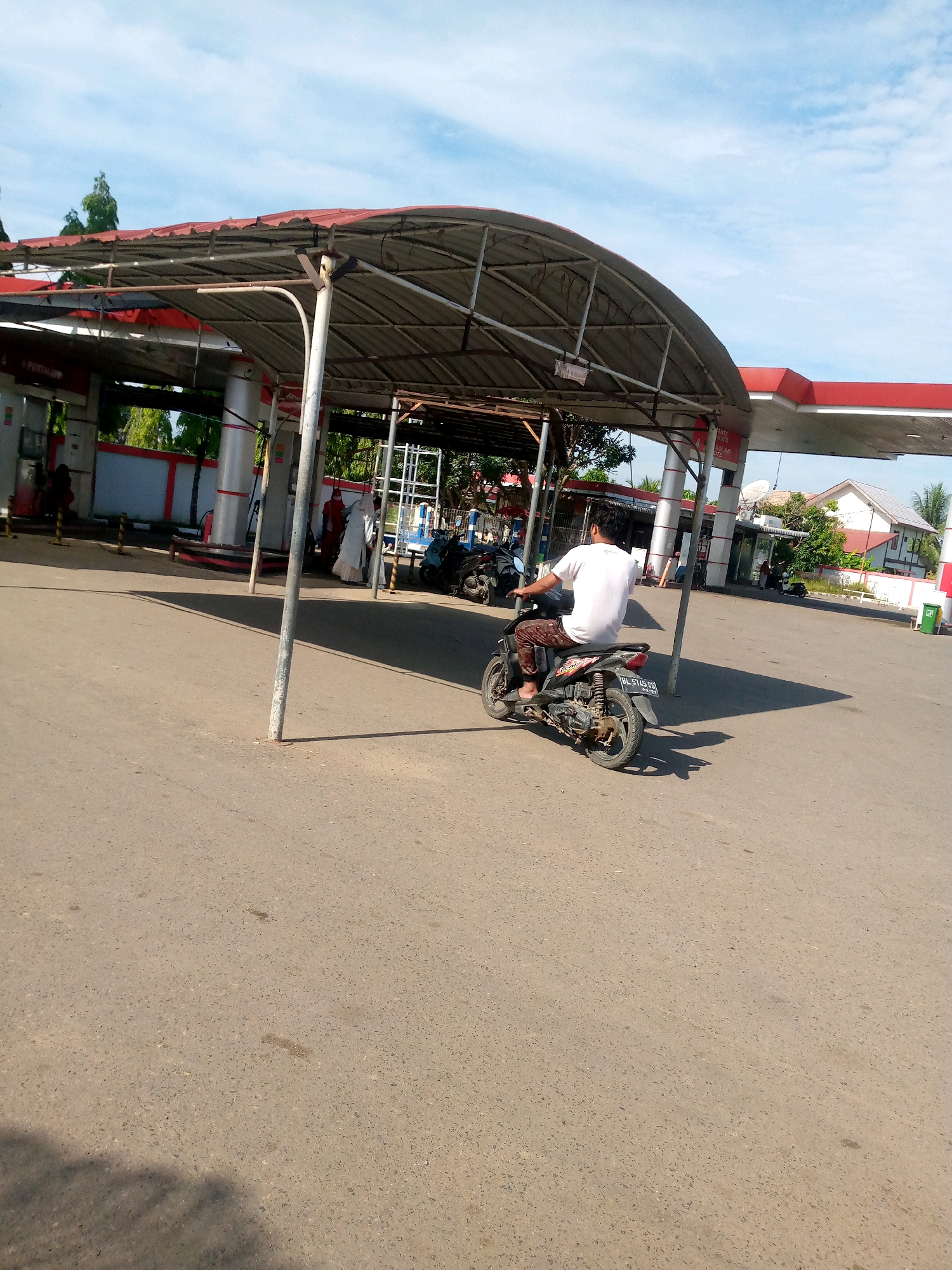 An outdoor scene at a gas station, identified as being in Keudebayu, Indonesia. The sun is shining, casting shadows. A man wearing a white t-shirt and patterned pants is riding a black motorcycle. The gas station features a red and white covered canopy supported by metal poles. In the background, there are gas pumps, buildings, trees, and more motorcycles. The ground is paved asphalt.
