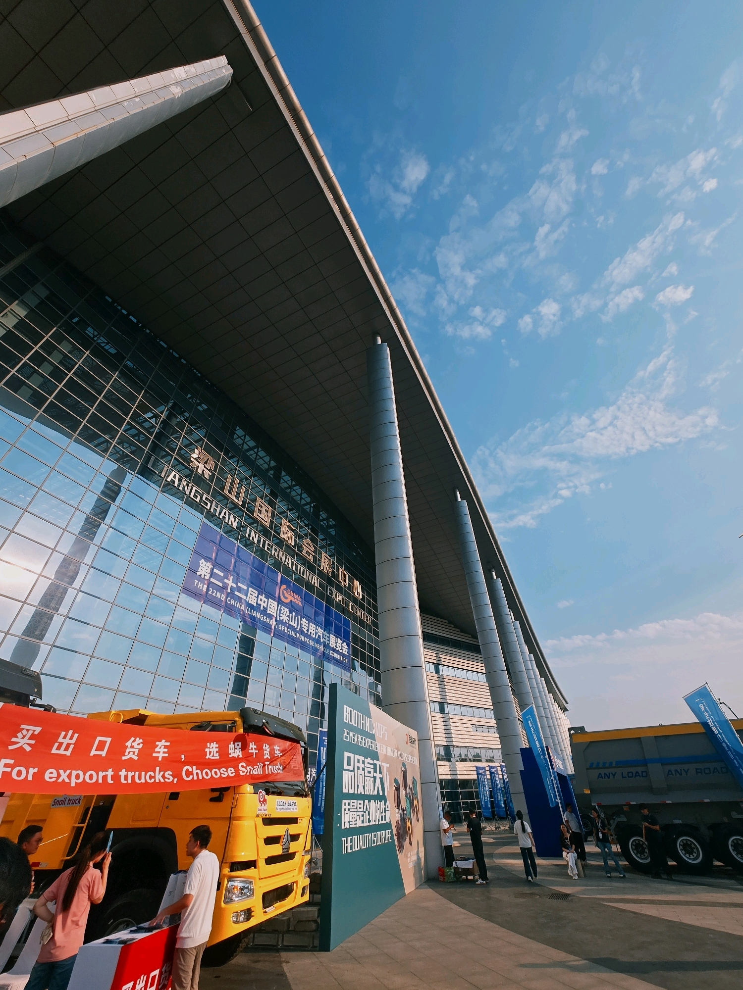 The image captures a sunny outdoor scene in front of the Liangshan International Expo Center in Liangshan County, China. The modern expo center features a large, glass-fronted facade and a distinctive overhanging roof supported by numerous tall, cylindrical pillars. In the foreground, a bright yellow truck is prominently displayed with a red banner that reads "For export trucks, Choose Snail Truck." Several individuals are interacting with the truck and nearby display booths. To the right, another large truck, possibly a dump truck, is visible with "ANY LOAD ANY ROAD" written on its side. The sky above is a clear blue with scattered white clouds, indicating good weather. The overall scene suggests an exhibition or trade show related to vehicles.
