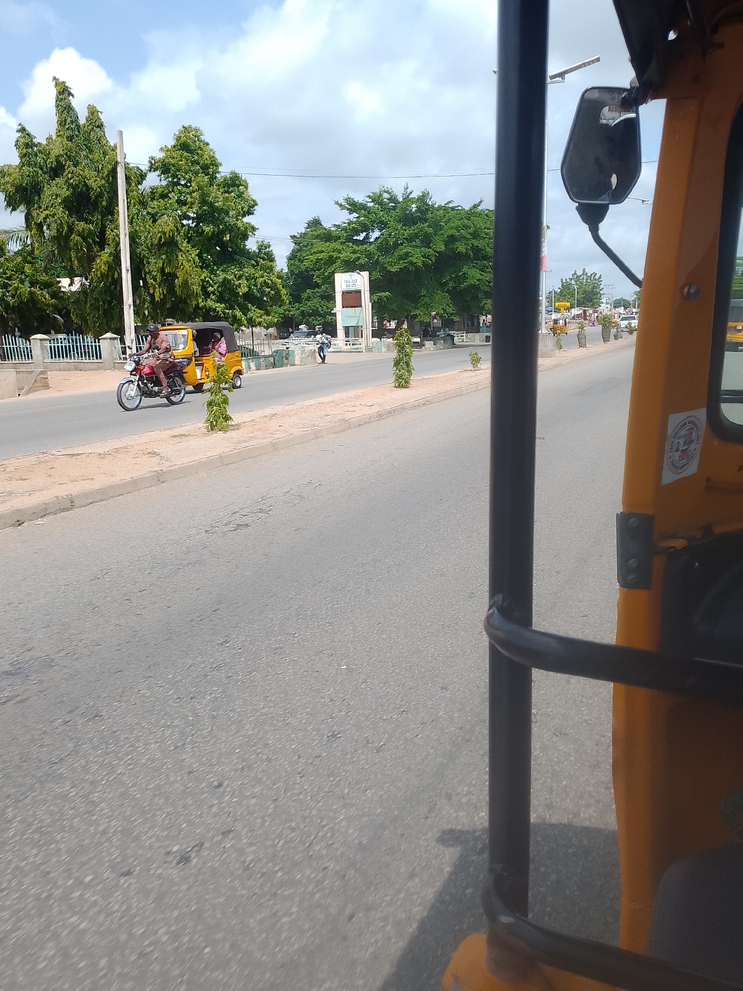 This media file shows a view from a moving yellow motorized tricycle, commonly known as a "Keke Napep" in Nigeria. The tricycle is on a paved road, likely in an urban or semi-urban setting, with a sidewalk and trees lining the road. The sky is partly cloudy, suggesting it is daytime.
In the foreground, part of the yellow tricycle's structure is visible, including a black metal bar and the side of the vehicle. The tricycle appears to be in motion, as indicated by the blur in the road and the perspective.
Further down the road, another tricycle is visible, carrying two passengers. The driver is wearing a brown shirt and pants, and the passengers appear to be a woman in pink and another person. The tricycle itself is yellow with a black canopy.
Other elements in the scene include tall green trees, a white pole, and a light post. In the distance, more trees, buildings, and other vehicles can be seen. A signpost with text is partially visible on the right side of the road in the distance. There's also a small sticker on the side of the tricycle in the foreground. The overall impression is a snapshot of daily life and transportation in a sunny, overcast day.