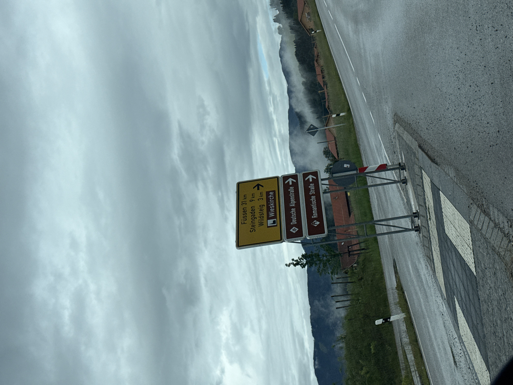 The image, taken in Rottenbuch, Germany, shows a roadside scene under an overcast sky. The primary subject is a multi-directional road sign in the foreground, indicating distances to various destinations and highlighting tourist routes.
The top, larger yellow sign displays destinations and their distances: "Füssen 31 km," "Steingaden 9 km," "Wildsteig 3 km," and "Wieskirche" with a church icon, indicating a right turn. Below this, two brown signs point right, identifying "Deutsche Alpenstraße" (German Alpine Road) and "Romantische Straße" (Romantic Road), both represented by a white diamond icon on a brown background.
To the right of the signs, a wet asphalt road stretches into the distance, marked with a white dashed line. A sidewalk with a cobblestone pattern is visible on the immediate right. In the middle ground, lush green grass separates the road from a row of wooden fences or barriers, behind which forested hills and mountains rise. A thick layer of fog or mist partially obscures the mountains, adding a serene or slightly melancholic atmosphere to the scene. The sky is entirely covered with thick, grey clouds, suggesting an overcast day, possibly after rain, as indicated by the wet road surfaces. No people or vehicles are visible.