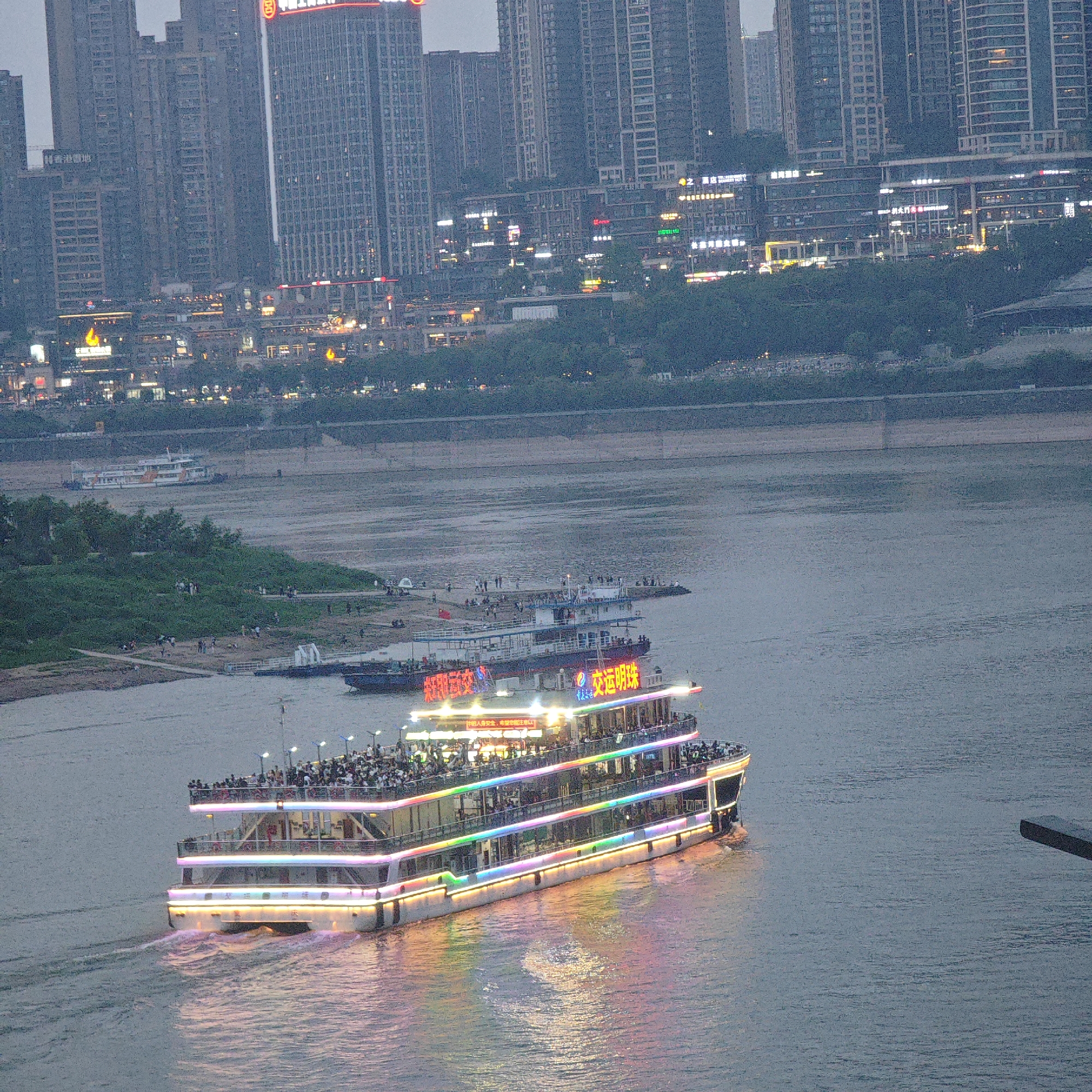The image depicts a river scene at dusk in Yuzhong District, Chongqing, China, showcasing a brightly lit multi-deck tour boat sailing on the river. The boat, illuminated with colorful lights, carries passengers. In the background, the city skyline is visible with numerous tall buildings and illuminated signs. The river water reflects the city lights. There are other vessels on the river and a small island with people. The overall environment suggests an urban setting at twilight.