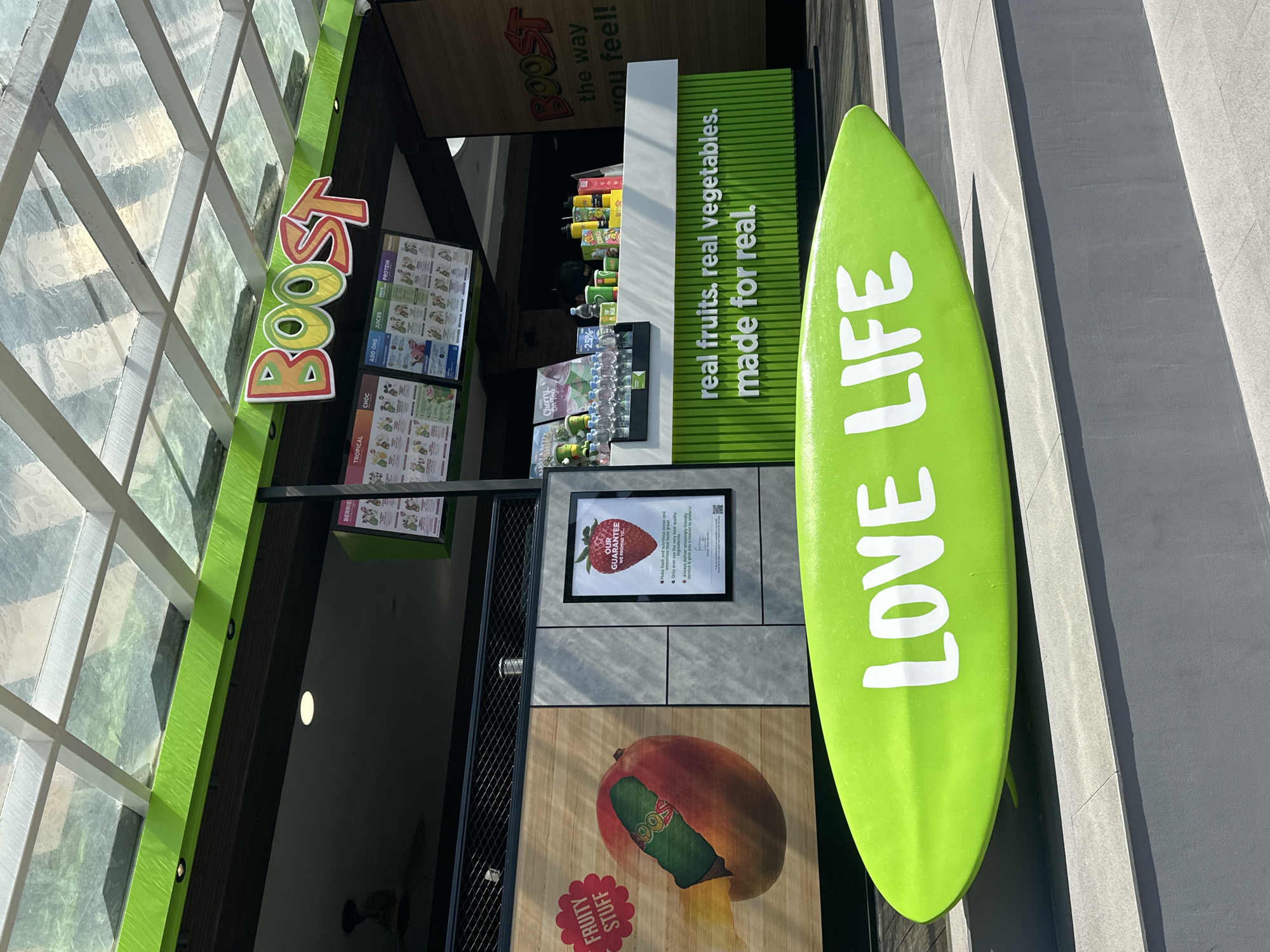 This image depicts a Boost Juice Bar storefront located in North Kuta, Indonesia. The store facade features a prominent lime green banner with the "BOOST" logo in multi-colored, bubble-style letters. Above the banner, a translucent glass roof with white structural beams is visible. Below the banner, several digital menu screens display various drink categories, including "TROPICAL," "CHOC," "ADD ONS," "JUICES," "CREATE," and "PROTEIN."
To the left of the counter, a large wall graphic shows a mango with a green, tubular object emerging from its side, accompanied by the text "FRUITY STUFF" in a red, starburst shape. Adjacent to this graphic is a framed sign titled "OUR GUARANTEE" featuring a strawberry image and legible text outlining quality promises, including fresh ingredients, friendly service, and a QR code. A black metal mesh barrier runs above these wall elements.
The counter area is primarily lime green and white, with the text "real fruits. real vegetables. made for real." inscribed in white on the green base. The counter holds various bottles, cups, and packaged items. To the far right, a vertical sign reads "BOOST the way you feel!" A dark interior is visible behind the counter, with a single ceiling light and a dark ceiling fan partially discernable.
In the foreground, a large, bright lime green surfboard-shaped display object rests on a grey tiled platform. The words "LOVE LIFE" are inscribed in white capital letters on its upper surface. The flooring in front of the store consists of multi-tiered grey tiles. Bright daylight illuminates the scene, casting distinct shadows. No identifiable people are present in the image.