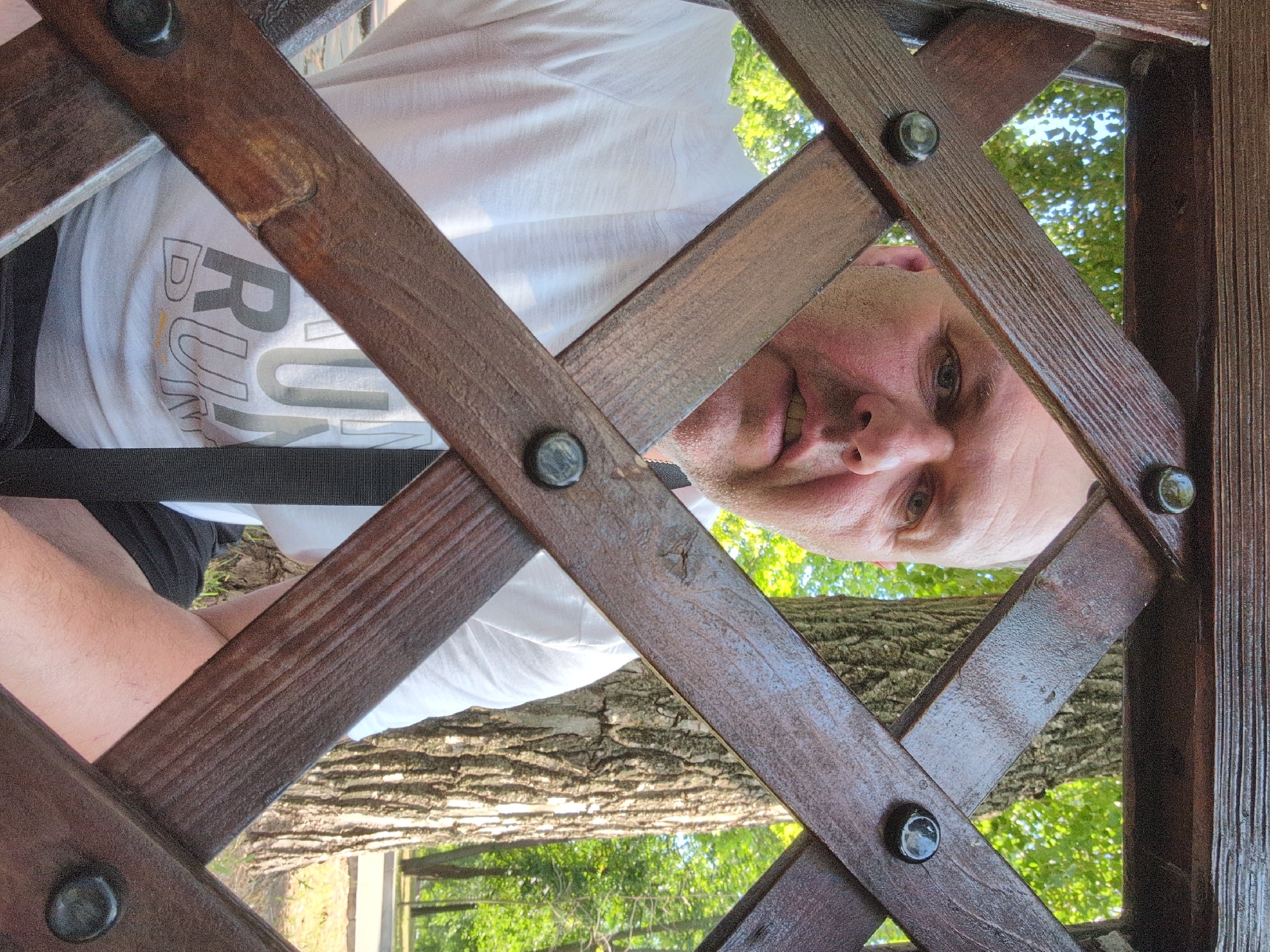 The image shows a man's face and torso looking up through a wooden lattice structure. The man is wearing a white t-shirt with the word "RUN" visible in large gray lettering. He appears to be outdoors, with trees and green foliage visible in the background, suggesting a park or natural setting. The lighting indicates it is daytime, likely sunny. The wooden lattice is dark brown and appears weathered, with visible screws or bolts holding the pieces together. The man's expression is neutral. The image is taken from a low angle, looking upwards. The location context is Kryvyi Rih, Ukraine, but there are no specific visual cues in the image to confirm this.