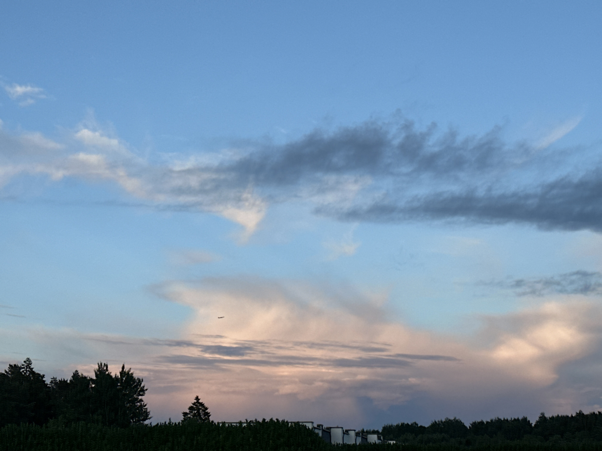 The image shows a twilight sky over Tiskre, Estonia. The sky is a blend of blue and pink hues, with scattered clouds. A small airplane is visible in the upper portion of the frame. Below the sky, the dark silhouettes of trees and a line of white cylindrical structures, possibly silos or storage tanks, are visible along the horizon. The overall scene is serene and captures the transition from day to night.