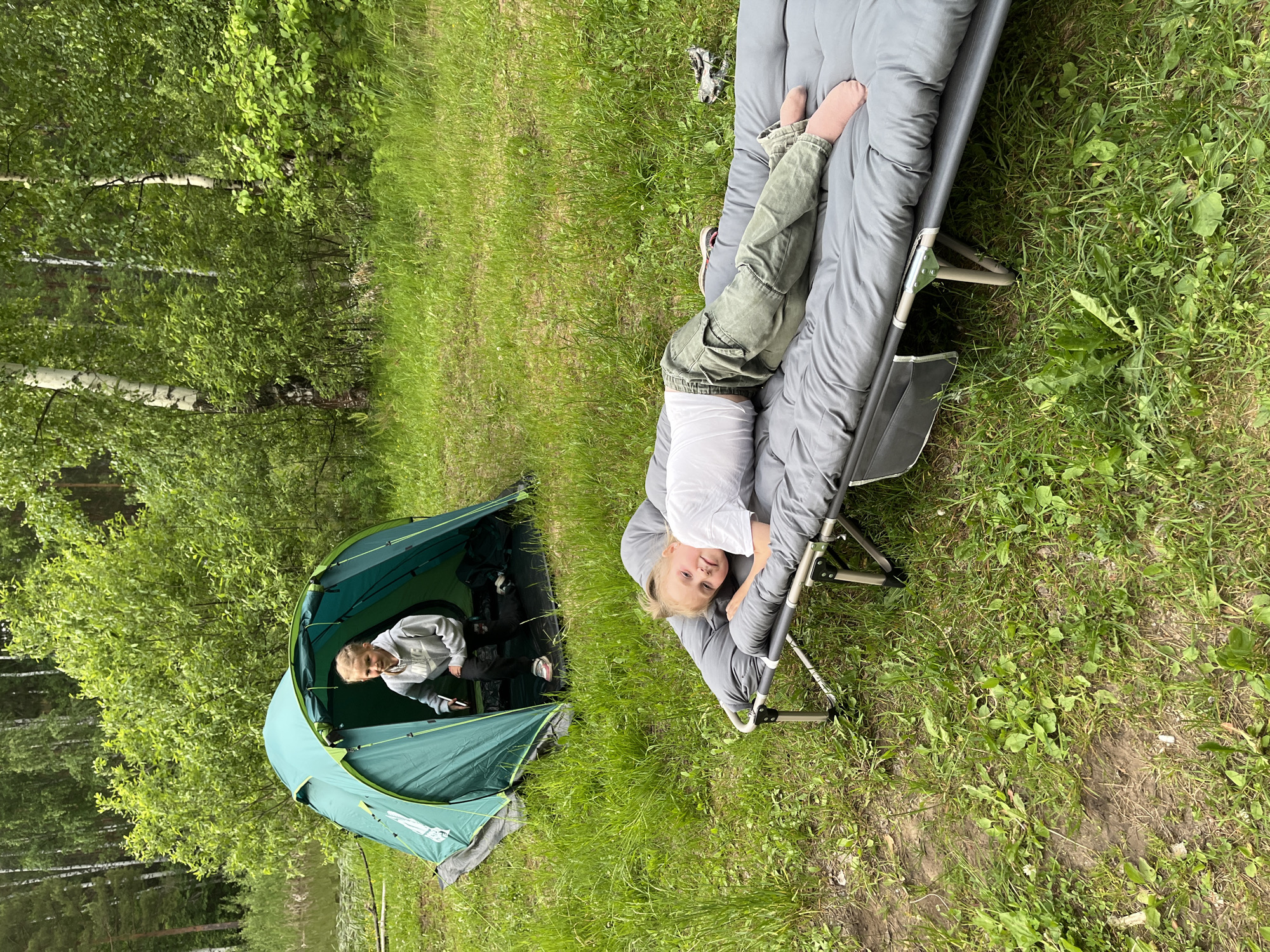 Two young children are outdoors near a lake and a forest. In the foreground, one child, wearing a white t-shirt and green cargo pants, lies on their side on a gray camping cot. In the background, a second child, wearing a gray hoodie and black pants, crouches inside a green tent. The scene is set in a grassy area with trees and foliage visible. The image appears to be taken during daylight hours.