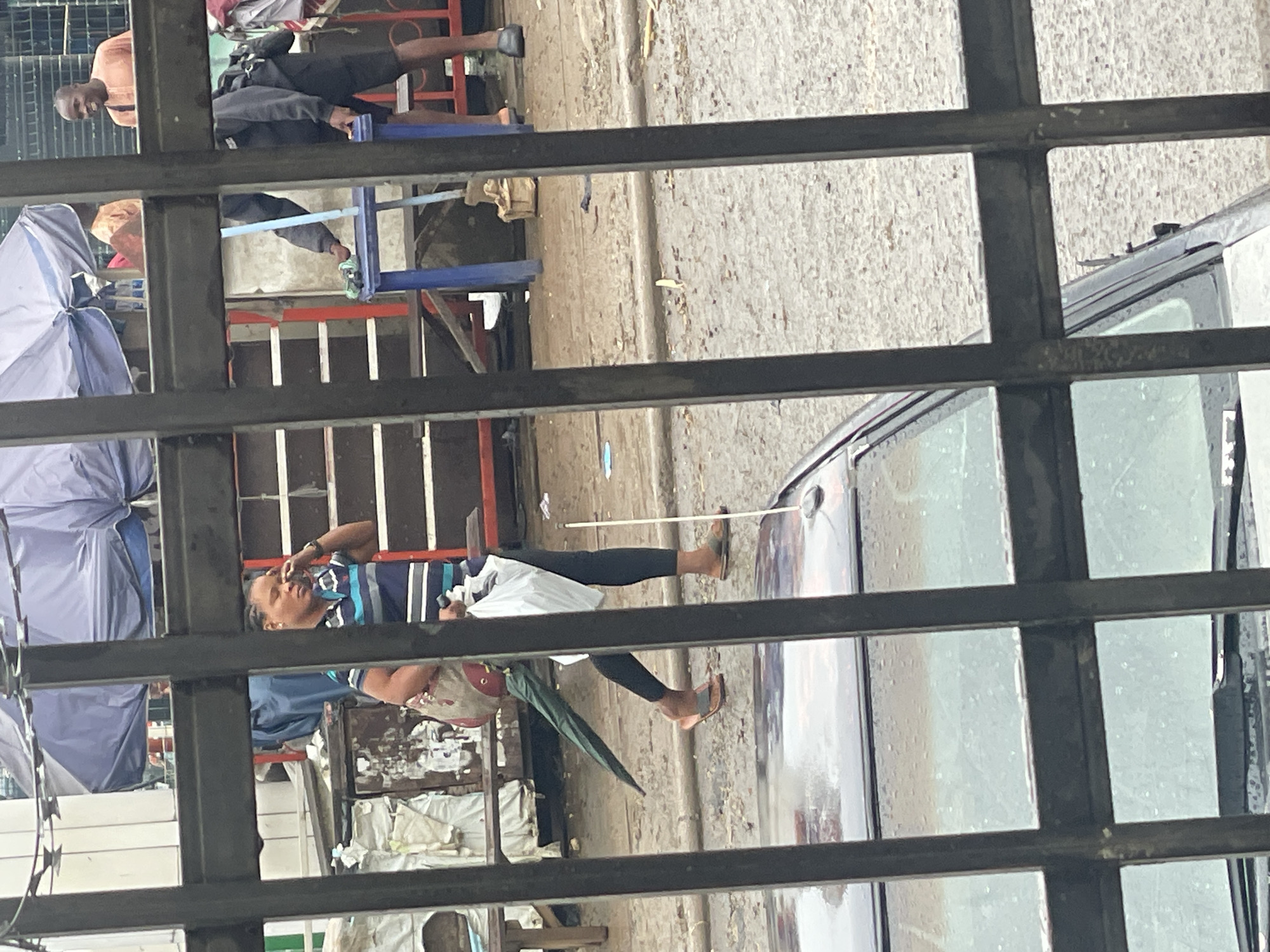 The image shows a street scene viewed through a metal fence. In the foreground, the roof and windshield of a dark-colored car are visible. The street surface is wet and appears to be asphalt with some debris.
In the mid-ground, a woman with braided hair is walking to the left, holding a white plastic bag in her right hand and a folded green umbrella in her left hand, tucked under her arm. She is wearing a horizontally striped shirt with blue, white, and dark blue colors, and black leggings. She is also wearing brown flip-flops. She appears to be talking on a mobile phone held to her left ear.
Behind the woman, there are some market stalls or makeshift structures with blue and white tarpaulin coverings. A blue stool or table is visible to the right of the woman. Further back, another person wearing an orange top is visible under a stall, and a man in a light orange shirt is smiling and looking towards the left. Another person in dark clothing and shorts is standing near the blue stool. In the background, there are more structures and what appears to be a fence or wall.
The scene suggests a busy outdoor market or street setting, possibly during or after rain, given the wet ground. The presence of market stalls and people engaged in various activities is typical of a commercial area. The overall impression is of a candid shot capturing a moment in daily life in an urban environment. This scene is consistent with a location in Abuja, Nigeria, which has numerous such street markets and commercial areas.
