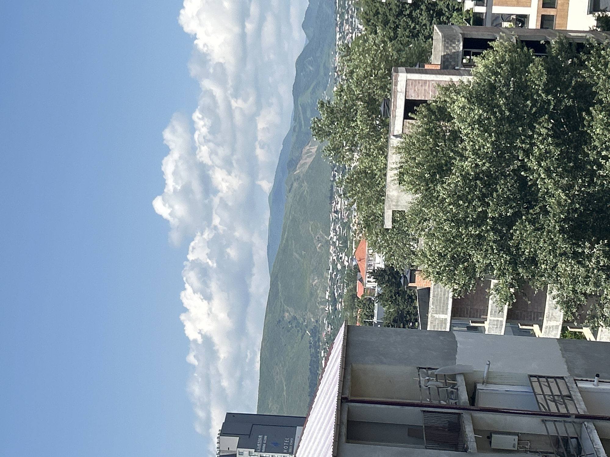 The image captures a view of a city nestled at the base of rolling green hills under a bright blue sky dotted with white cumulus clouds. In the foreground, a partially constructed building with exposed concrete and brickwork is visible. To the left, a grey high-rise building displays the word "HOTEL" and some text in Georgian script. A section of another multi-story apartment building with balconies is also present on the far left. A dense canopy of green trees occupies the middle ground, partially obscuring a town with red-tiled roofs and other buildings. The distant hills are covered in lush vegetation. The overall impression is of an urban area surrounded by natural landscape on a clear day.