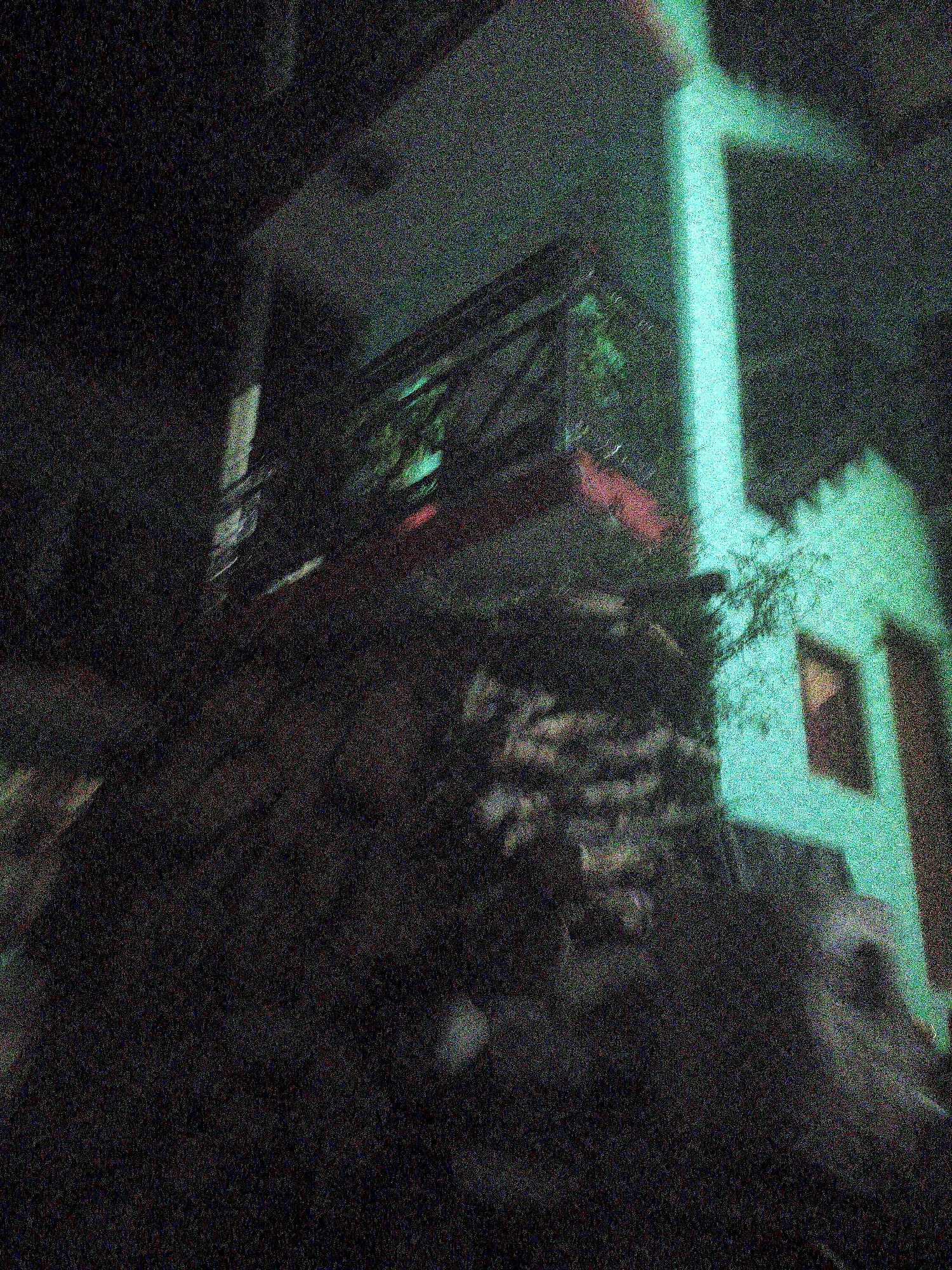 The image depicts a dimly lit, outdoor scene at night, with a significant amount of digital noise present, obscuring finer details. In the foreground, a dark, textured surface, possibly rough stone or earth, dominates the lower portion of the frame. Above this, a structure with a balcony is visible. The balcony features a railing, and within its confines, what appear to be plants or foliage can be discerned. The building behind the balcony is illuminated with a strong, unnatural green light. This green light highlights structural elements of the building, including a vertical beam and a section of wall with a dark, recessed window or opening. A smaller, lighter-colored structure is partially visible to the right, also illuminated by the green light. The overall impression is of a nighttime view of a building, possibly residential or commercial, in an urban or semi-urban environment, with artificial green lighting casting a dramatic effect on the scene. Given the context of Vashisht, India, this could be a view of a building in the town, potentially near a hot spring or a temple, given the prevalence of such lights in religious and tourist areas.
