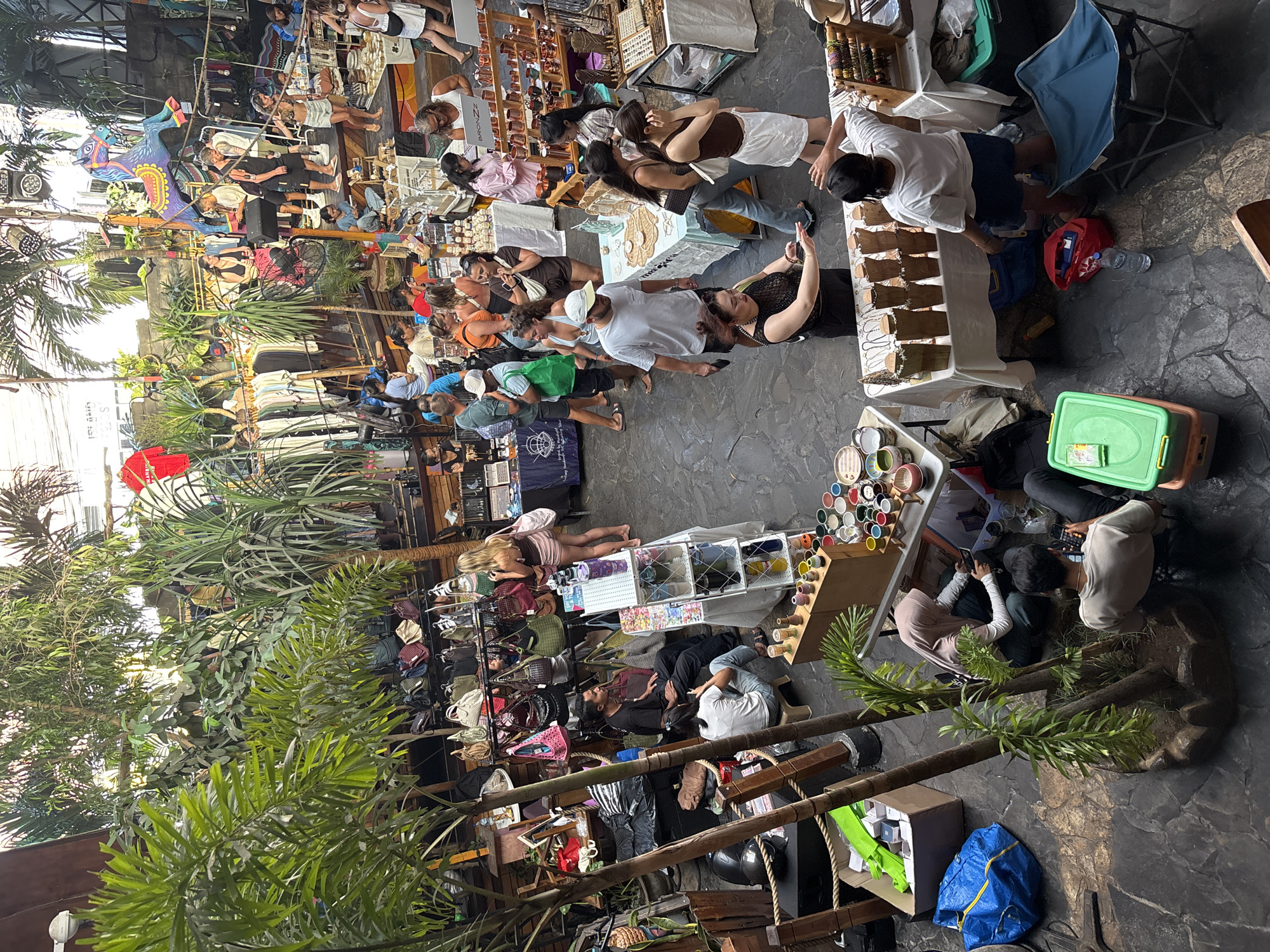 This aerial view captures a vibrant indoor market in Kuta Selatan, Indonesia, filled with people browsing various stalls. The market is lush with tropical plants, including palm trees, creating a bohemian atmosphere. Stalls display an array of goods such as handbags, jewelry, clothing, and decorative items. Several people are seen interacting with vendors, examining merchandise, and congregating in groups. In the foreground, a blue IKEA bag and a green plastic container are visible on the stone floor. The overall scene depicts a busy and lively marketplace.