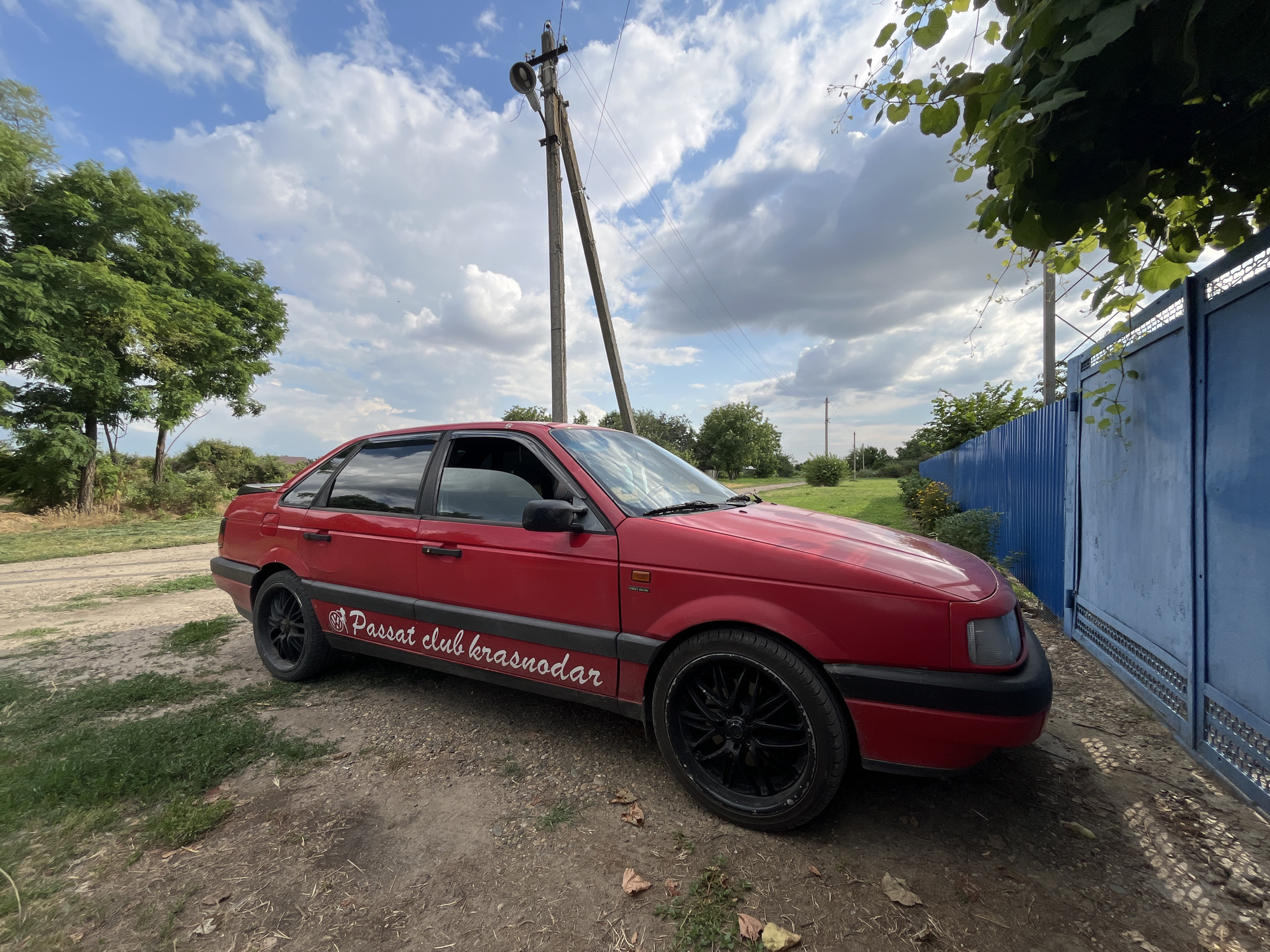 A red Volkswagen Passat sedan is parked on a dirt road next to a blue corrugated metal fence and gate. The car has a sticker on its side that reads "Passat club krasnodar." A utility pole with a lamp is visible behind the car, with power lines extending across the frame. Trees and green foliage are present on the left side of the image, and a larger tree with dense leaves overhangs the top right corner. The sky is cloudy with patches of blue. The scene appears to be in a rural or semi-rural setting, possibly the village of Otrado-Olginskoye, Russia, given the context of the sticker.