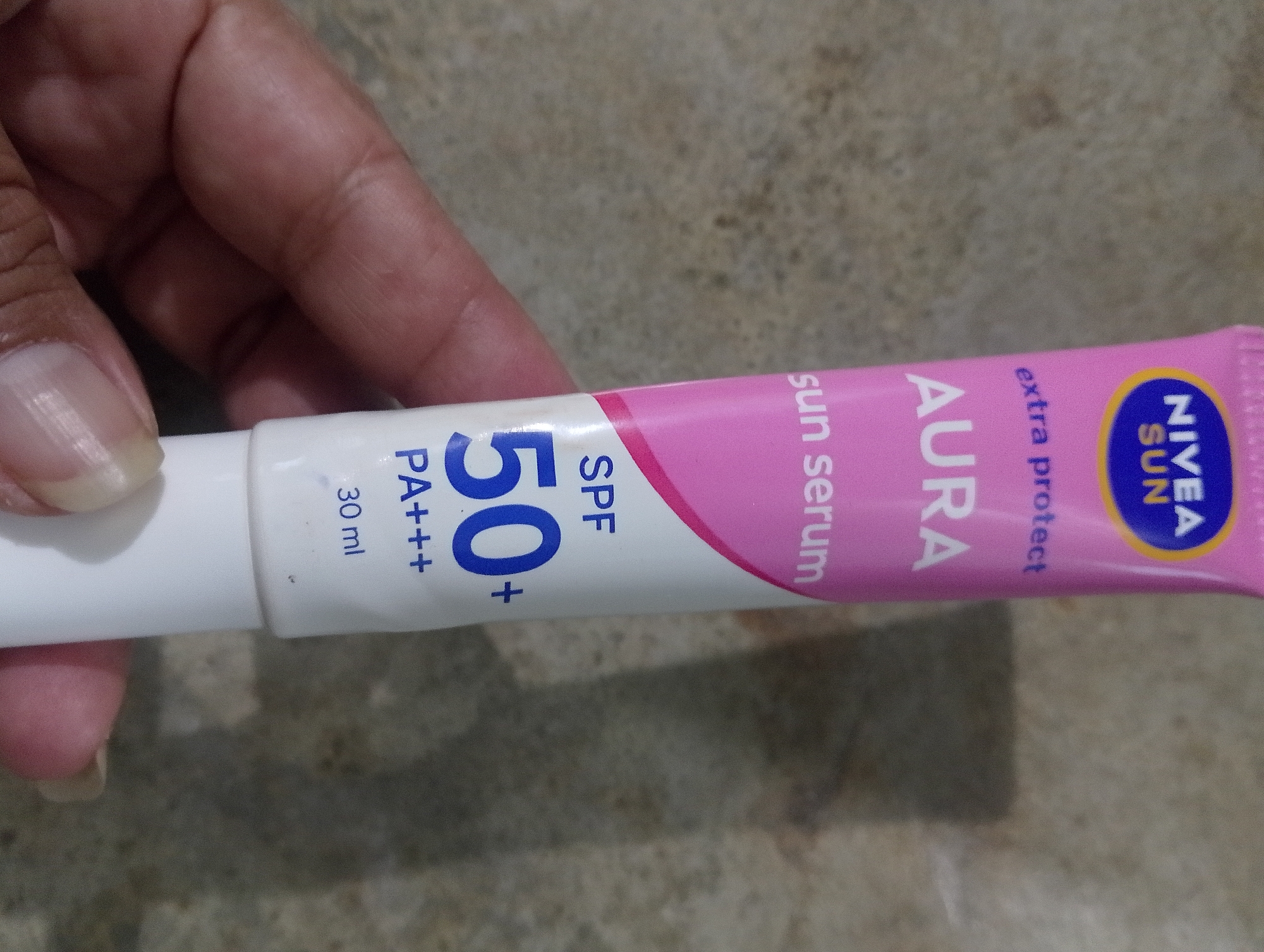 A hand holds a white and pink tube of Nivea Sun Aura SPF 50+ PA+++ Sun Serum, 30 ml. The tube has blue and yellow Nivea Sun branding on the right side. The background appears to be a mottled brown surface, possibly a countertop or floor, and the image is cropped to focus on the product and the hand. The hand's fingers are visible gripping the tube. The location of Serbelawan, Indonesia, is not depicted in the image.