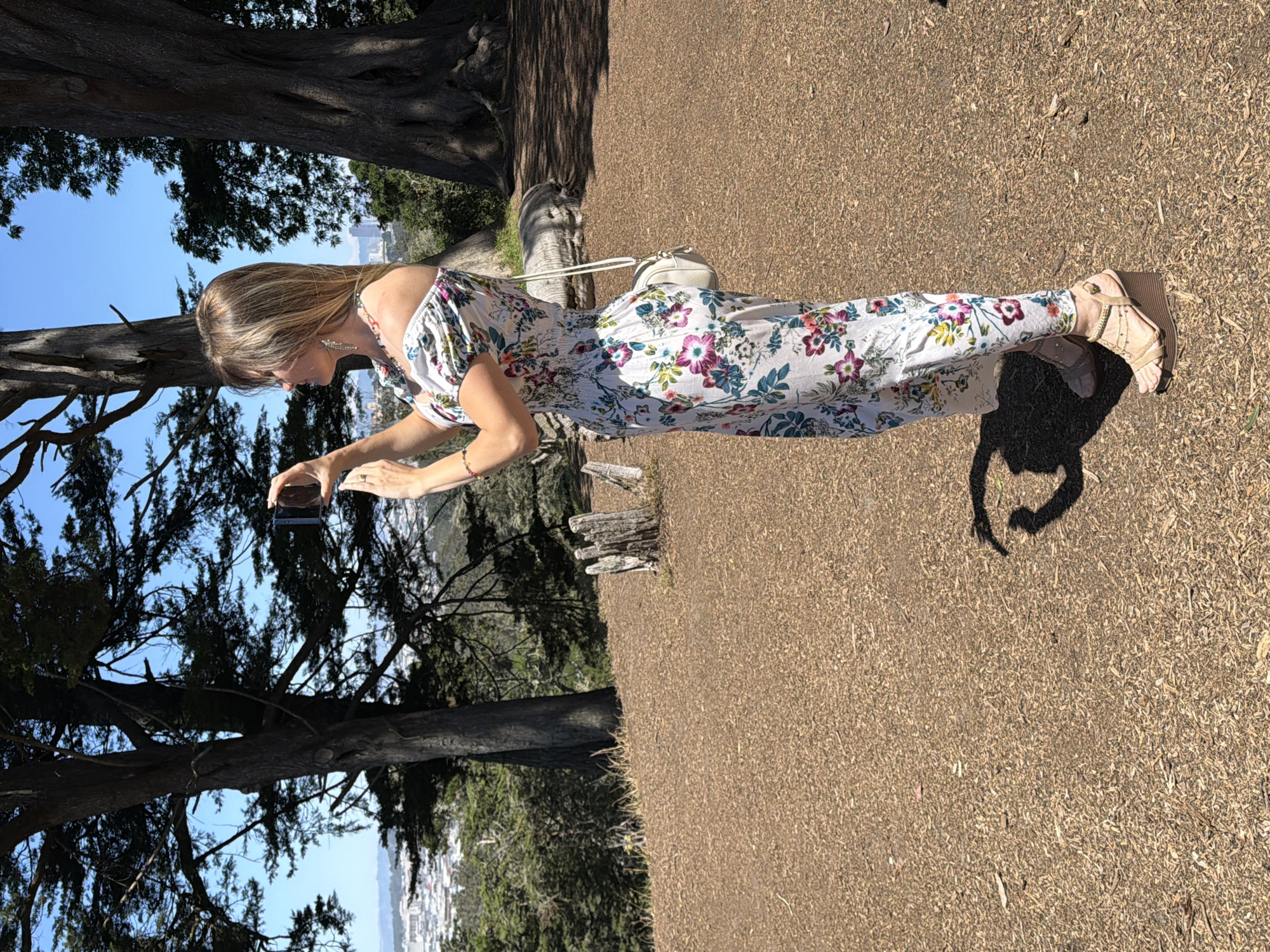 A Caucasian woman with blonde hair is standing on a dirt path overlooking a city. She is wearing a white off-the-shoulder dress with a floral print, a white crossbody bag, and wedge sandals. She is holding a smartphone in her hands and appears to be taking a photo. The background shows trees and buildings, indicating the location is San Francisco. The woman is facing away from the camera, towards the city. The scene is lit by the sun, casting a shadow of the woman on the ground.