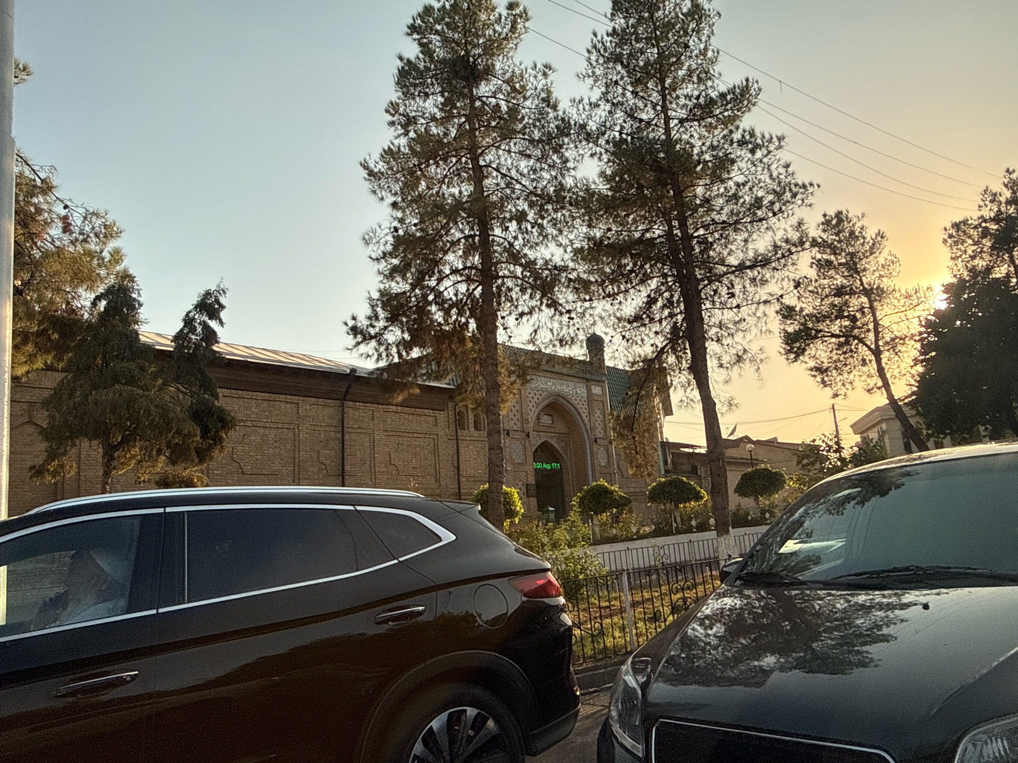 In Tashkent, Uzbekistan, during daylight hours, two dark-colored cars are parked on a street. The car on the left, a black SUV, is closer to the viewer and its rear section is visible. The car on the right, a dark sedan, is partially visible in the foreground. Behind the cars, a brick building with ornate architectural details, possibly a historical site or a mosque, is prominent. The building features arched entrances and intricate carvings. Several tall evergreen trees with thin trunks and sparse foliage stand in front of the building and along the street, some silhouetted against the bright, hazy sky. A metal fence runs along the sidewalk. The overall setting appears to be an urban environment with a blend of modern vehicles and traditional architecture, bathed in the warm light of what appears to be late afternoon or early morning sun.