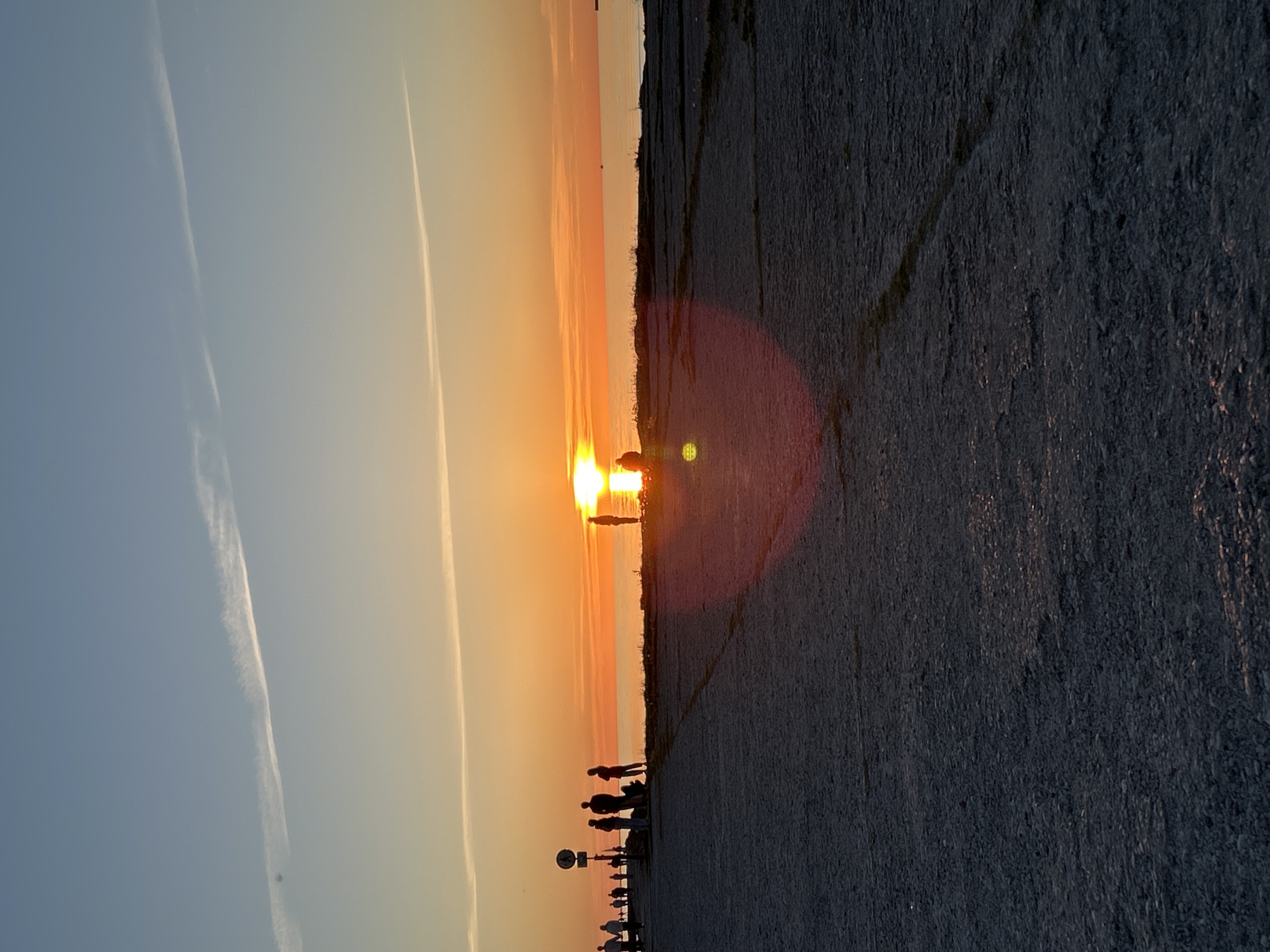 This image depicts a sunset over a calm body of water, identified as the Baltic Sea, from an urban waterfront promenade in Tallinn, Estonia. The sun, a brilliant orange-yellow orb, is positioned centrally on the horizon line, casting an intense light that transitions the sky from deep blue at the top to warm orange and yellow hues closer to the horizon. Two distinct, long white contrails traverse the upper sky diagonally from left to right.
The foreground consists of a dark, coarse aggregate paved surface, likely concrete or asphalt, occupying the lower two-thirds of the image. This surface is largely in shadow, exhibiting subtle warm reflections from the sunset, and shows linear patterns indicative of paving sections. A prominent circular lens flare artifact with warm tones is visible in the lower-middle foreground.
Numerous individuals are present as dark silhouettes, observing the sunset. On the left side, a group of approximately four people stands near a dark signpost on a slightly elevated paved section, with three additional figures positioned further left along the promenade. Two prominent figures stand at the water's edge, directly in the path of the sun's reflection, and one individual is seated on the ground in the mid-foreground. Sparse, dark vegetation is visible between the paved area and the water. The water surface is calm, reflecting a bright orange-yellow band from the sun's direct path.