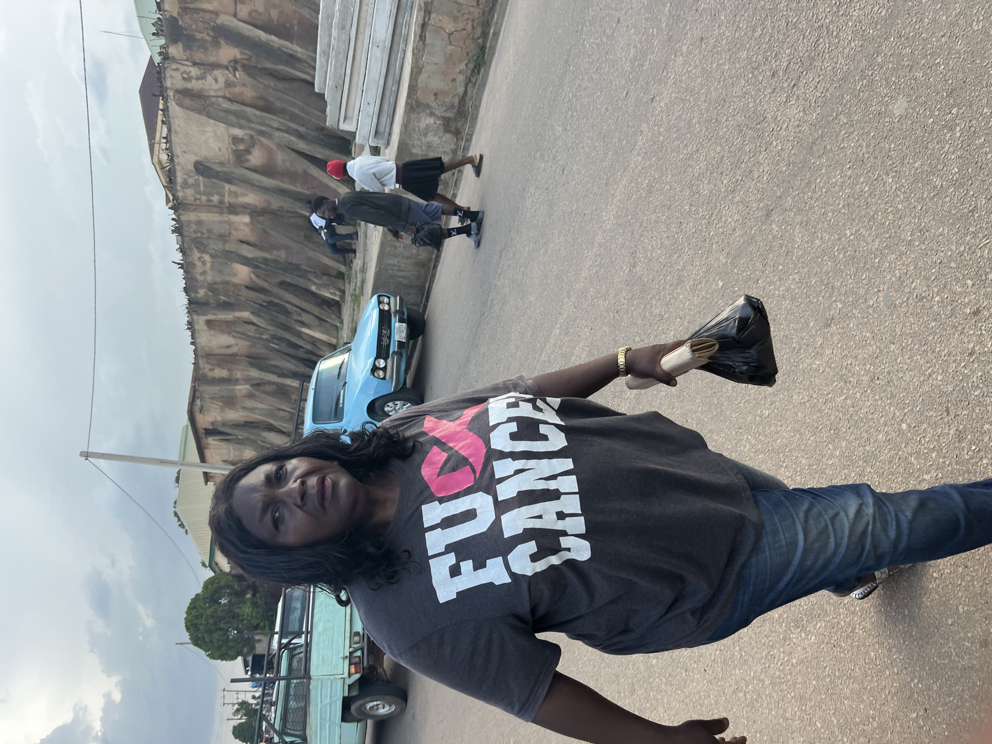 A medium shot captures a woman walking towards the camera on a paved road in Gura Top, Nigeria. She has dark, wavy hair and is wearing a charcoal grey t-shirt with "FUCK CANCER" printed in large white letters, with the "CK" of "FUCK" replaced by a pink ribbon graphic. She is also wearing blue jeans and sandals. In her right hand, she carries a black plastic bag. Behind her, a light blue car is parked on the right side of the road, and a light blue pickup truck is visible on the left. In the background, a large, rough-hewn concrete wall with angled supports forms a barrier. Two individuals, a man in a dark long-sleeved shirt and shorts and a person in a white long-sleeved shirt and a black skirt with a red beanie, are walking away from the camera on the right side of the road, near the wall. The sky is overcast with scattered clouds.