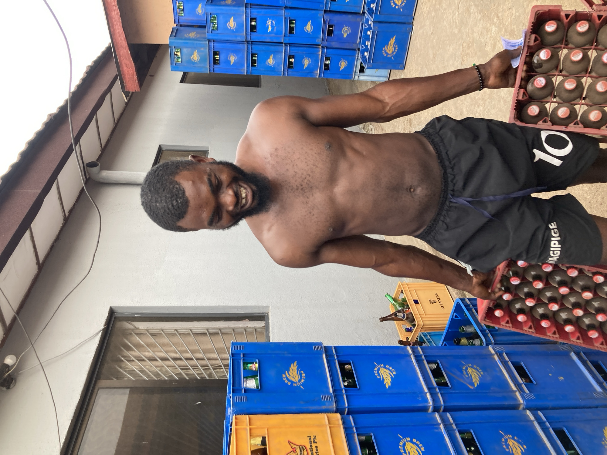 A muscular, shirtless man with a beard is standing in what appears to be a storage or warehouse area, possibly a beverage depot. He is holding two red crates filled with bottled drinks. The man is smiling, suggesting he is involved in the distribution or handling of the beverages. Behind him, there are numerous stacks of blue and yellow plastic crates. Some of the crates contain bottles, indicating they are likely for transporting drinks. The environment is enclosed by a gray building with a window and a gutter. The overall scene suggests a business or commercial context, likely in Jos, Nigeria given that "Premium Brew" which are stacked on the blue crates.