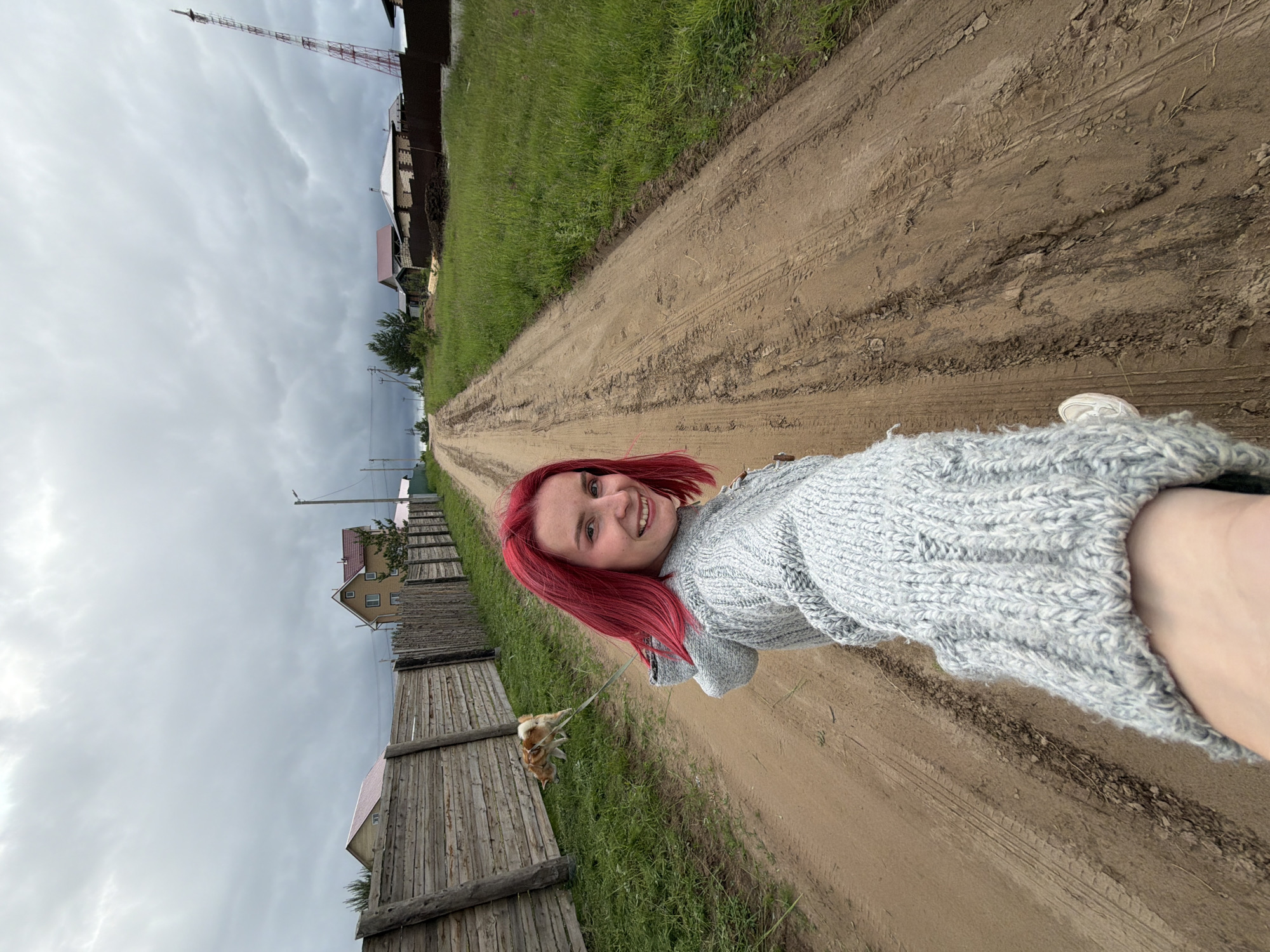 A young adult female with distinct bright red, shoulder-length hair is captured in a selfie-style photograph, smiling directly at the viewer. She extends her right arm, holding the camera, and wears a light grey, chunky knit cardigan. Her left foot, clad in a white sneaker, is visible in the lower left. She is walking a small to medium-sized dog, possibly a Shiba Inu or similar breed with tan and white fur, on a light-colored leash held by her left hand. The dog walks on the left side of a wide, unpaved dirt road, with its head lowered, appearing to sniff the ground.
The scene depicts a rural or semi-rural environment in Chaikovsky, Russia. The dirt road, marked with numerous tire tracks and footprints, extends uphill into the background, flanked by grassy verges. To the left, a rustic wooden fence constructed from horizontal planks and vertical posts runs parallel to the road. Beyond the fence and further down the road, several houses with reddish-brown and lighter-colored roofs are visible, interspersed with trees and utility poles. On the right side of the road, a wider strip of overgrown green grass and weeds leads to more houses, some appearing to be under construction (with exposed brickwork), and a taller, darker fence. A prominent red and white striped lattice communication tower stands on the far right horizon, extending high into the overcast, grey sky, which lacks any visible direct sunlight.
