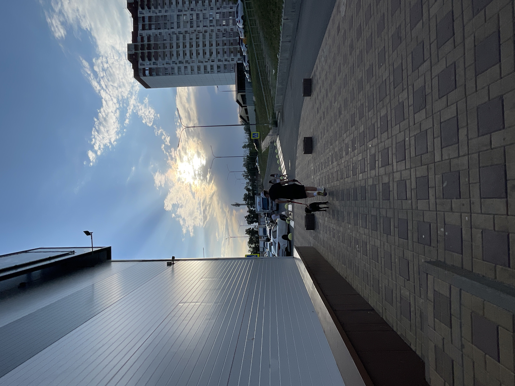 The image shows a paved walkway next to a white building with horizontal panels. A person in dark clothing is walking a black dog on a leash along the walkway. In the background, there is a street with cars, a tall apartment building, streetlights, and a cloudy sky with the sun partially visible, casting rays of light. The scene appears to be in a city environment, possibly Rostov-on-Don, Russia, given the architectural style of the apartment building and the overall urban setting. There are also some yellow bollards and a pedestrian crossing sign visible on the street. The pavement is made of interlocking rectangular and square stones.