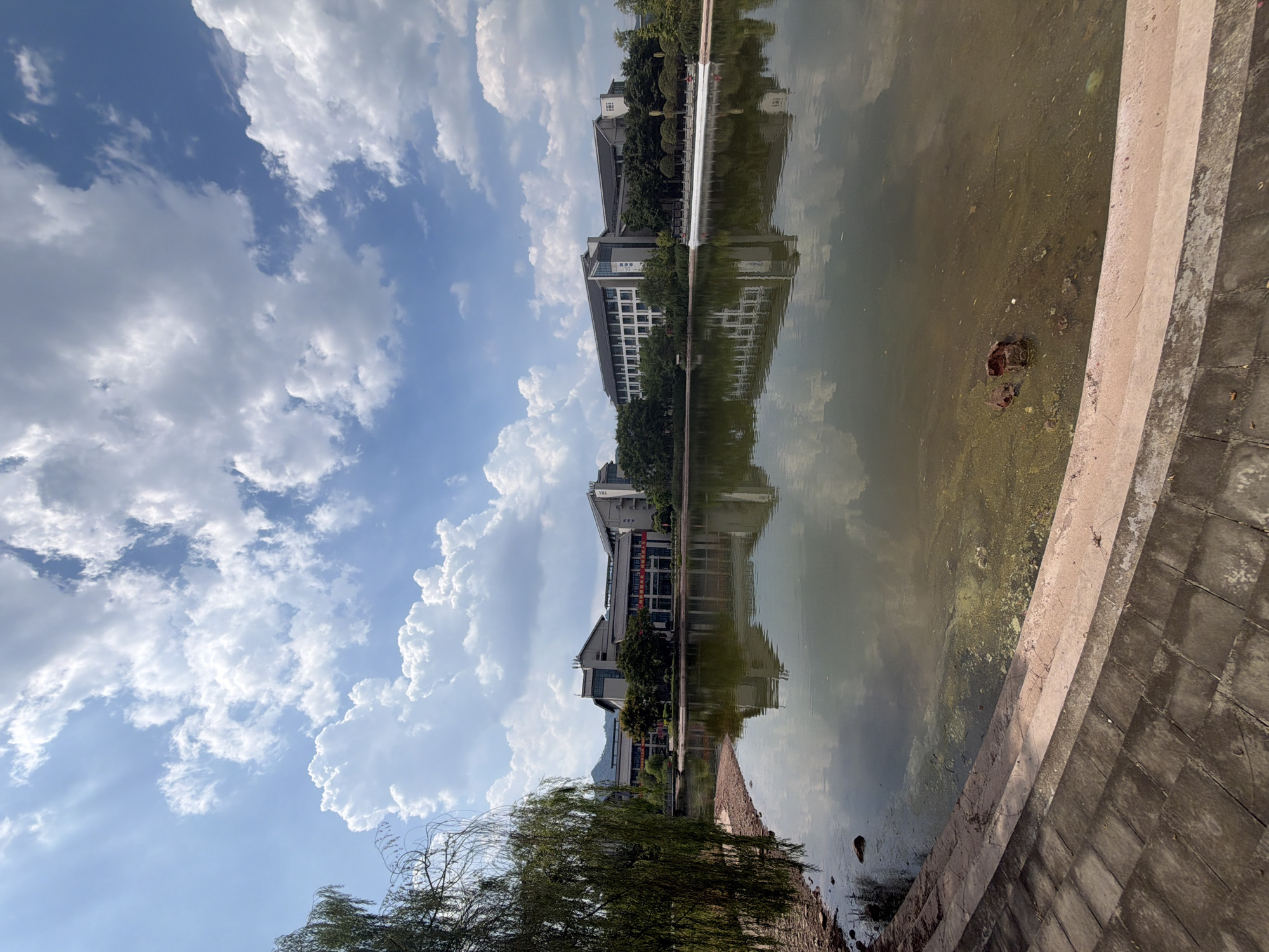 A wide-angle, eye-level shot captures a serene scene at the Jiangjin University campus in Jiangjin, China. The foreground features a stone-lined walkway leading to a calm lake. Lush green trees, including a weeping willow on the left, fringe the water's edge, their reflections mirroring in the placid surface. Across the lake, a modern, multi-story building complex stands, its architecture characterized by clean lines and dark-tiled roofs. The building is surrounded by a variety of trees. The sky above is a brilliant blue, dotted with fluffy white clouds that cast gentle shadows on the landscape. The overall impression is one of peaceful academic life within a picturesque natural setting.