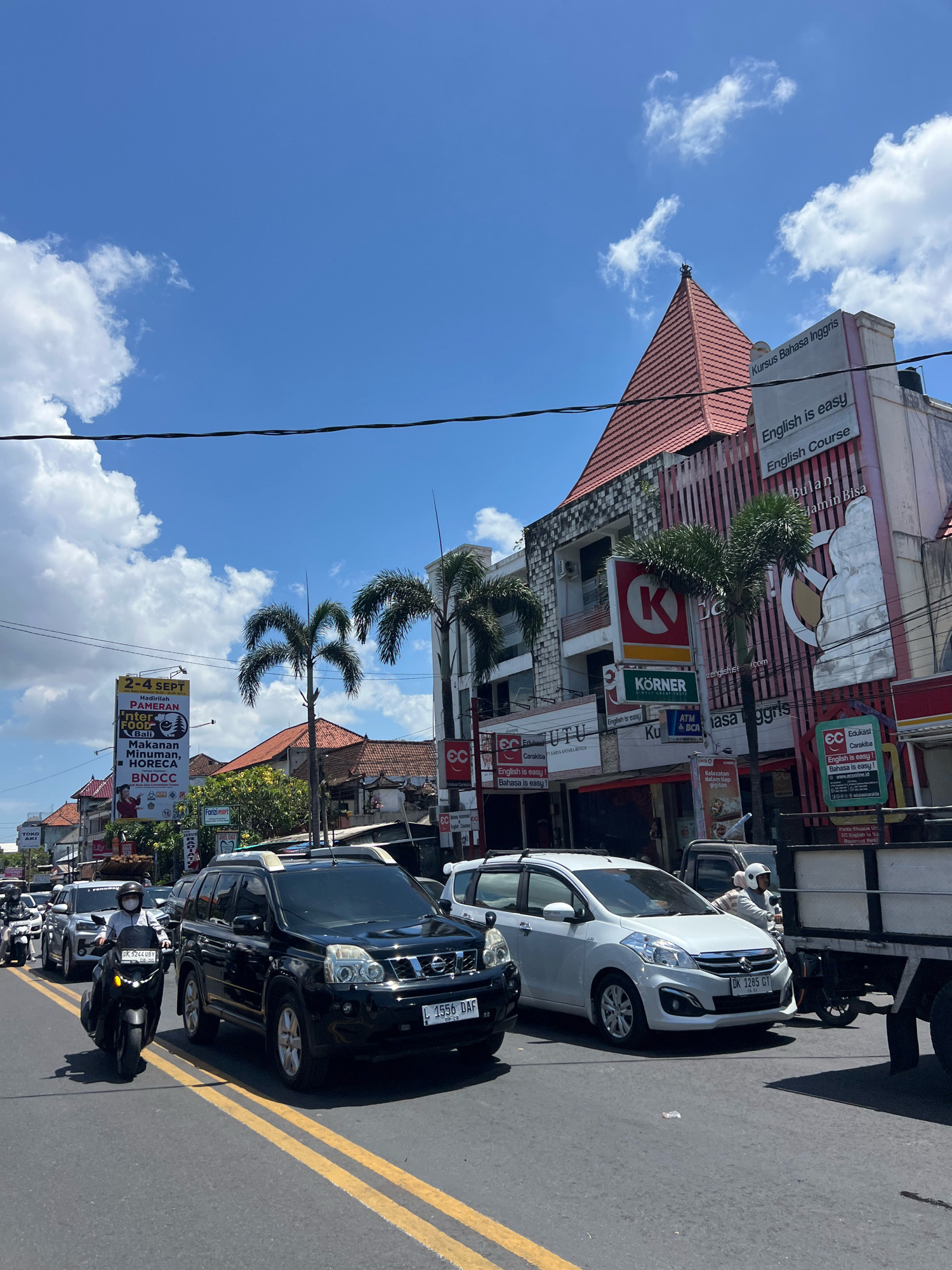 The image depicts a street scene in Denpasar, Indonesia, under a bright blue sky with scattered white clouds. The street is busy with traffic, including cars, motorcycles, and a truck. In the foreground, a black SUV and a white minivan are stopped in traffic, with yellow lines marking the lanes on the asphalt road. A motorcyclist wearing a helmet and mask is seen to the left, filtering through traffic.
The buildings lining the street feature a mix of architectural styles, with some exhibiting traditional Balinese elements such as red tiled roofs. Prominent signage indicates businesses, including an "English is easy" language course, a "Körner" sign, and a "Funzii mart" advertisement. Other signs are in Indonesian.
The overall impression is a typical urban streetscape in a tropical climate, characterized by vibrant activity and commercial establishments.