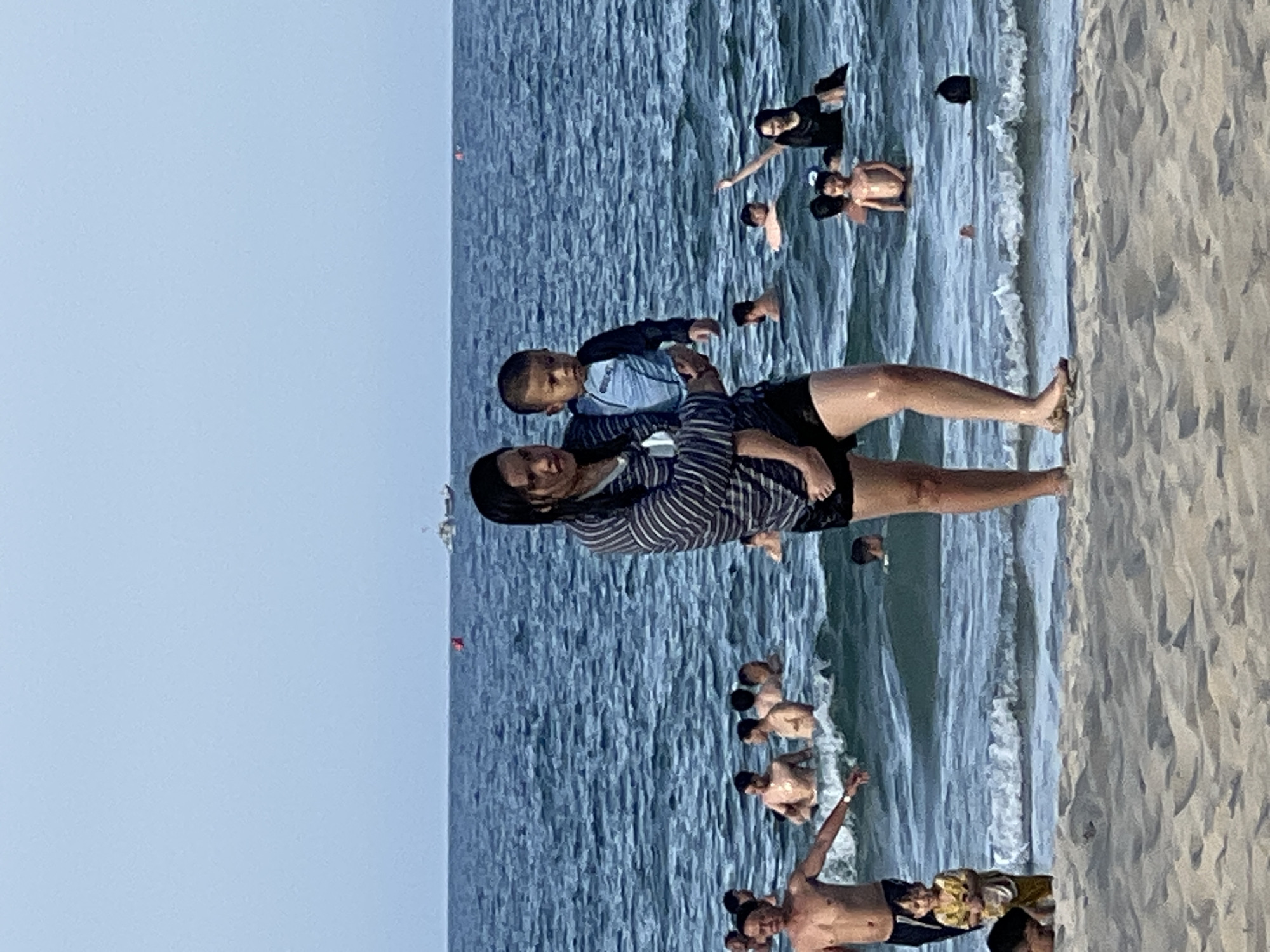 A medium shot captures a beach scene in Da Nang, Vietnam, with a woman holding a toddler in her arms as they stand at the edge of the water. The woman is wearing a striped long-sleeved shirt and black shorts, and the toddler is dressed in a blue and black rash guard. Numerous other people are visible in the ocean, wading, swimming, and playing in the waves. In the distance, a boat is visible on the horizon under a clear blue sky. The foreground shows a sandy beach with gentle waves lapping at the shore.