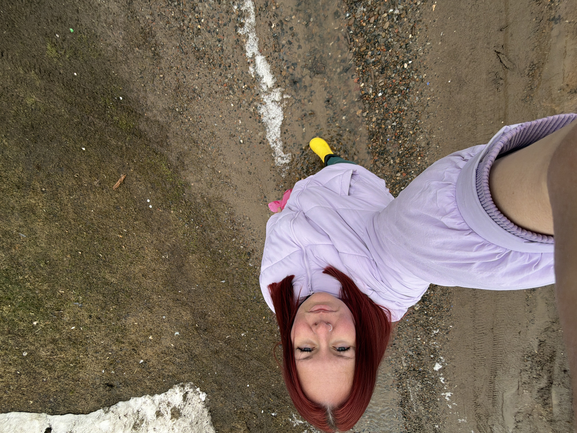 A woman with red hair and a nose ring takes a selfie-style photograph while standing outdoors. She is wearing a light purple jacket and yellow rubber boots, and a small amount of snow can be seen. The ground appears to be wet. According to geolocation, the photograph was taken in Chaikovsky, Russia.