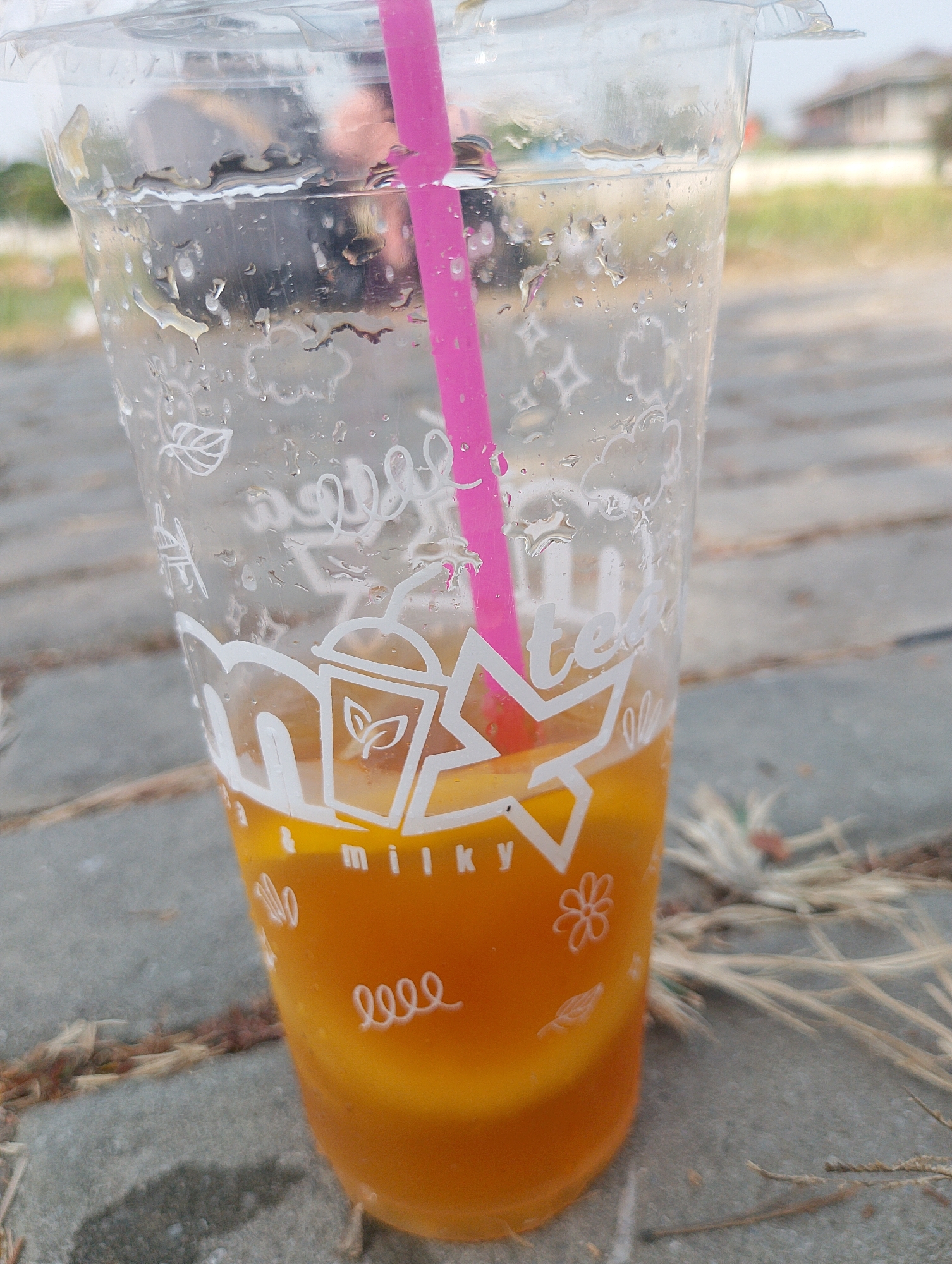 A close-up, eye-level shot captures a clear plastic cup filled with an orange beverage and ice. A pink straw extends from the top of the cup. The exterior of the cup is adorned with white printed designs, including stars, leaves, flowers, and text that partially reads "milky" and "tea." Condensation is visible on the outside of the cup, indicating it's a cold drink. The cup sits on a paved surface, possibly a sidewalk or patio, composed of gray rectangular stones. In the background, out of focus, is a blurry expanse of green grass and some structures, suggesting an outdoor setting in Karawang, Indonesia. Dried grass stalks are scattered on the paving stones around the cup.
