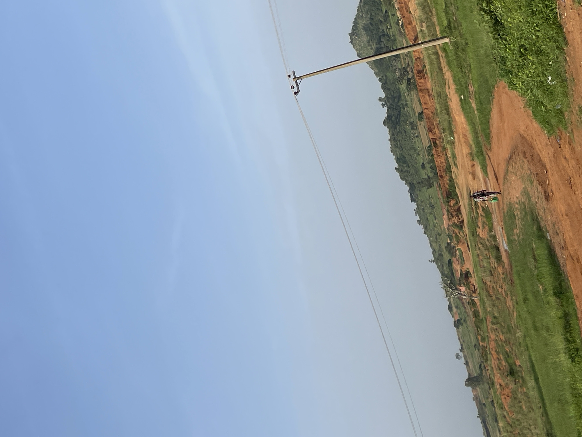 This image depicts an outdoor scene in Barikin Tin Tin, Nigeria, likely during daylight hours given the bright sky. The photograph is oriented vertically, with the sky occupying the left two-thirds of the frame and a landscape filling the right third. A utility pole with wires stretches diagonally across the image from the upper left to the lower right, indicating infrastructure presence.
The landscape features a dirt road winding through green, grassy fields and barren, reddish-brown earth that suggests terra firma. The terrain appears slightly elevated in parts, with trees and vegetation dotting the hillsides. In the distance, a motorcyclist with a passenger is visible on the road, suggesting travel or transport. A few white specks, possibly birds, are scattered across the grassy areas. The overall atmosphere is serene and rural, typical of a countryside setting. There is no visible text in the image.