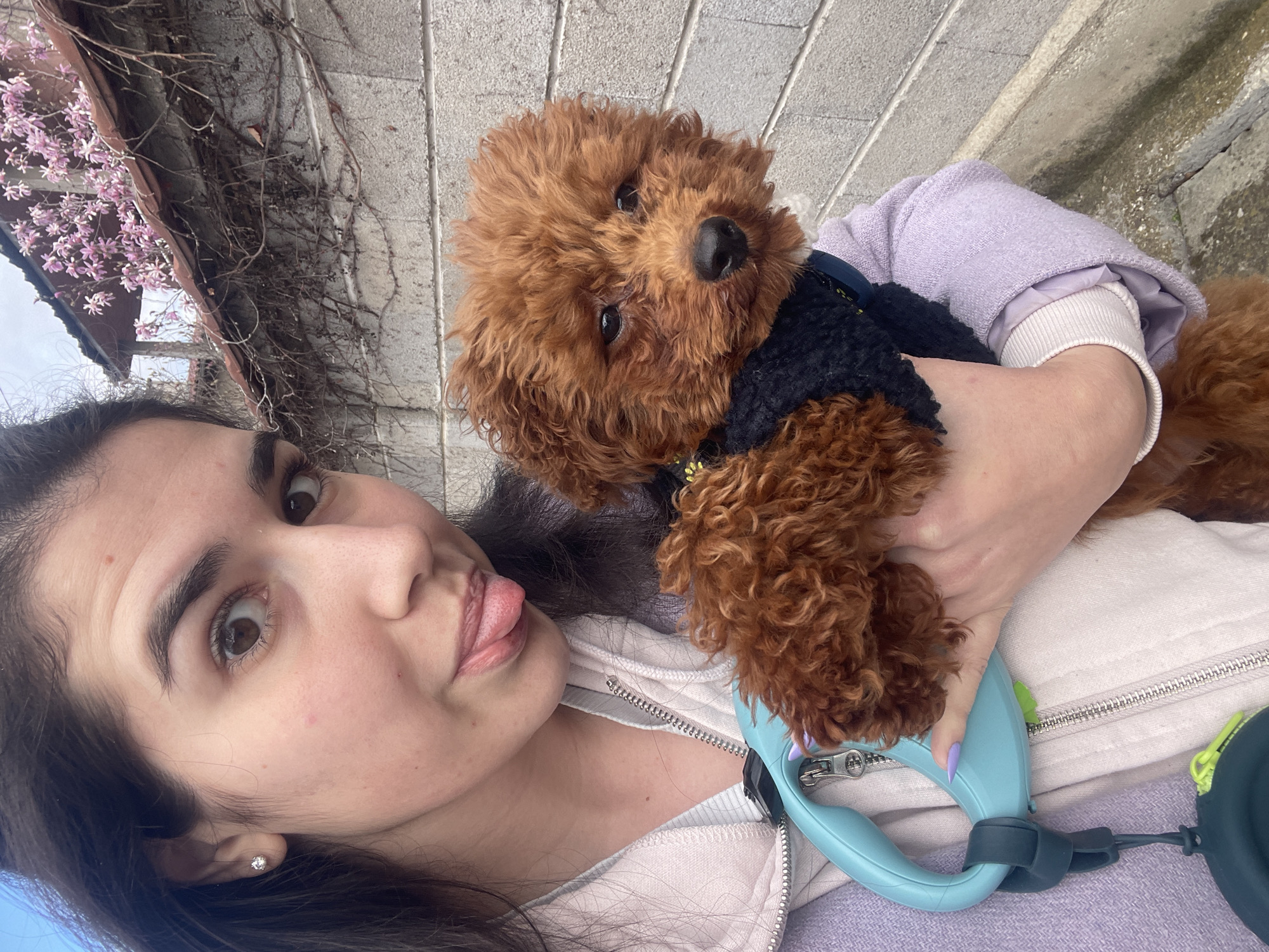 A young woman is taking a selfie with a small, brown poodle. She is holding the dog close to her chest. The woman has dark hair and light skin. She is wearing a light-colored hoodie and a light purple jacket. The dog is wearing a black and navy blue jacket. A blue dog leash is visible. In the background, there is a building with climbing vines and pink flowers. The location is likely Sofia, Bulgaria, based on visual cues typical of the region.