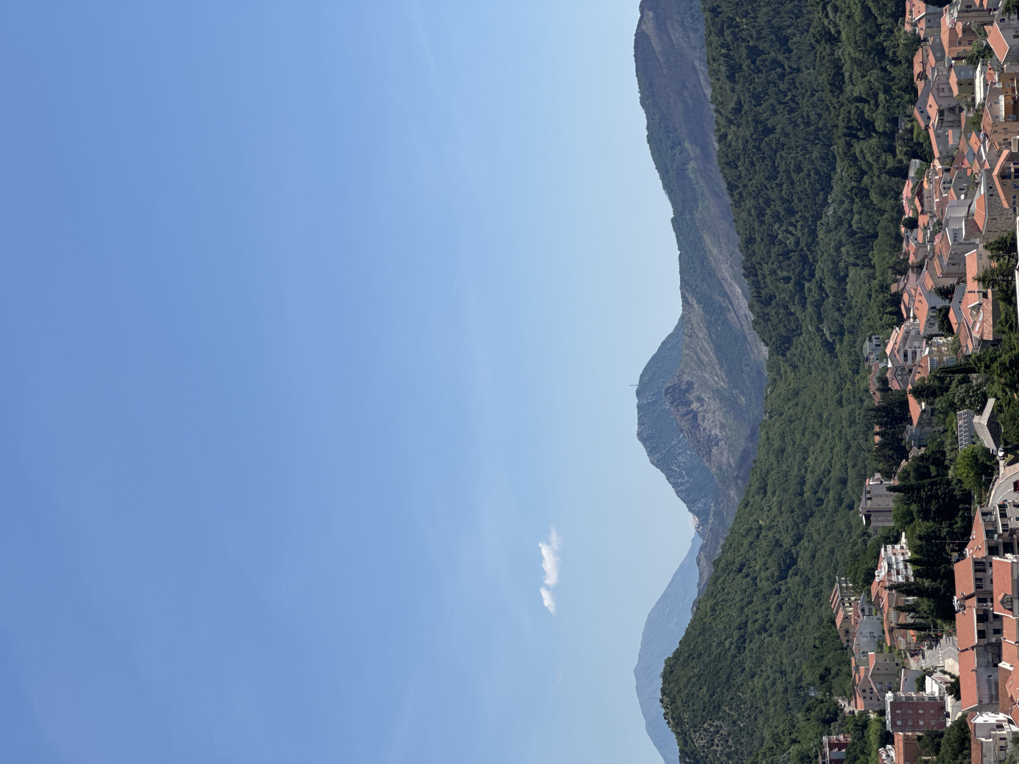 The image presents a view in Nerin, Montenegro, showcasing a mountain range densely covered with green trees. The terrain slopes down to a cluster of buildings with red roofs indicating residential or commercial structures. A clear blue sky dominates the background.