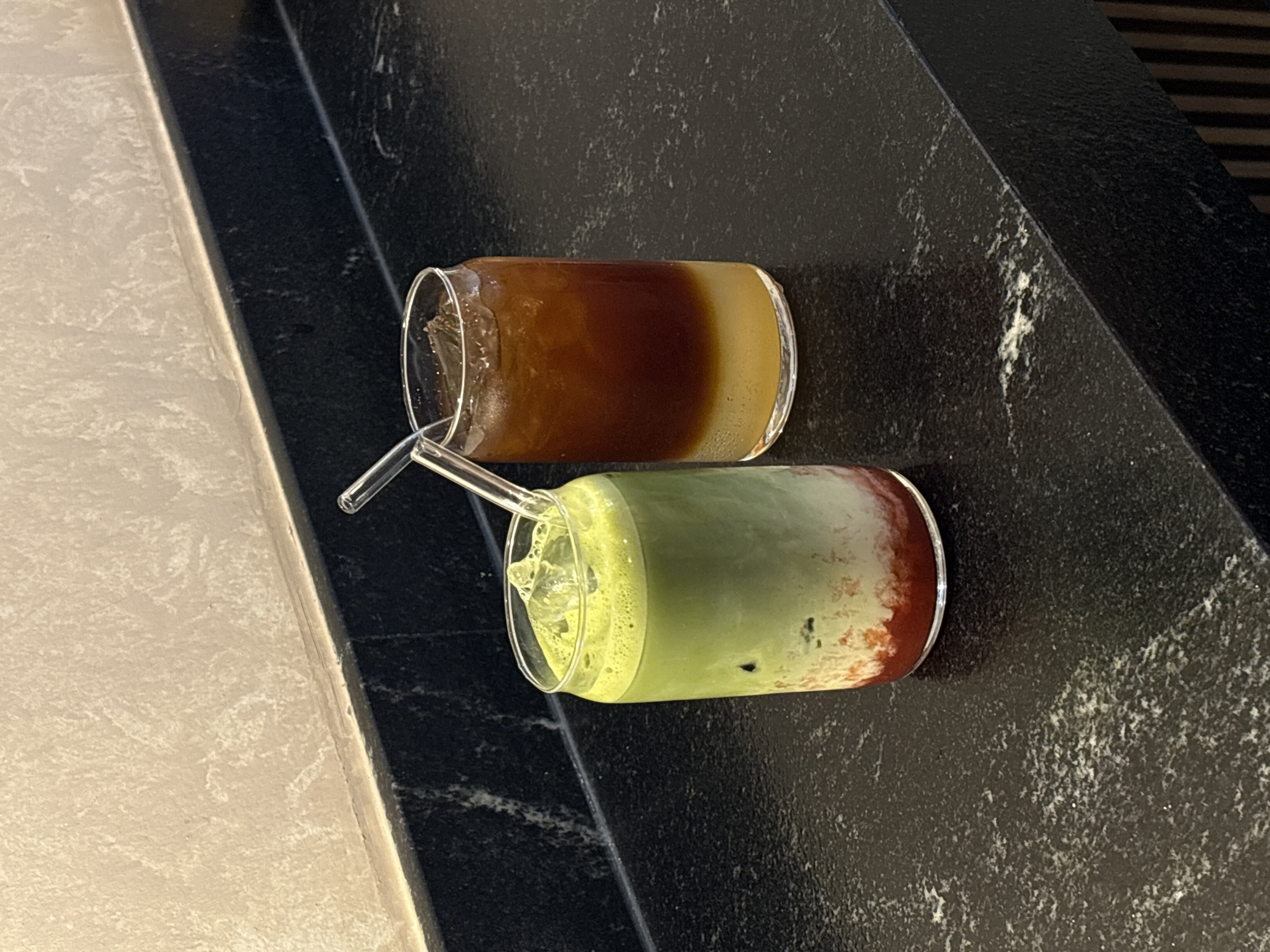 Two drinks in glass cans, each with a bent glass straw, are placed on a dark, textured countertop. One drink is layered brown and yellow, while the other is layered green, yellow, and red, with ice visible. The countertop edge is adjacent to a light-colored wall or counter. The location is San Bernardino Tlaxcalancingo, Mexico.