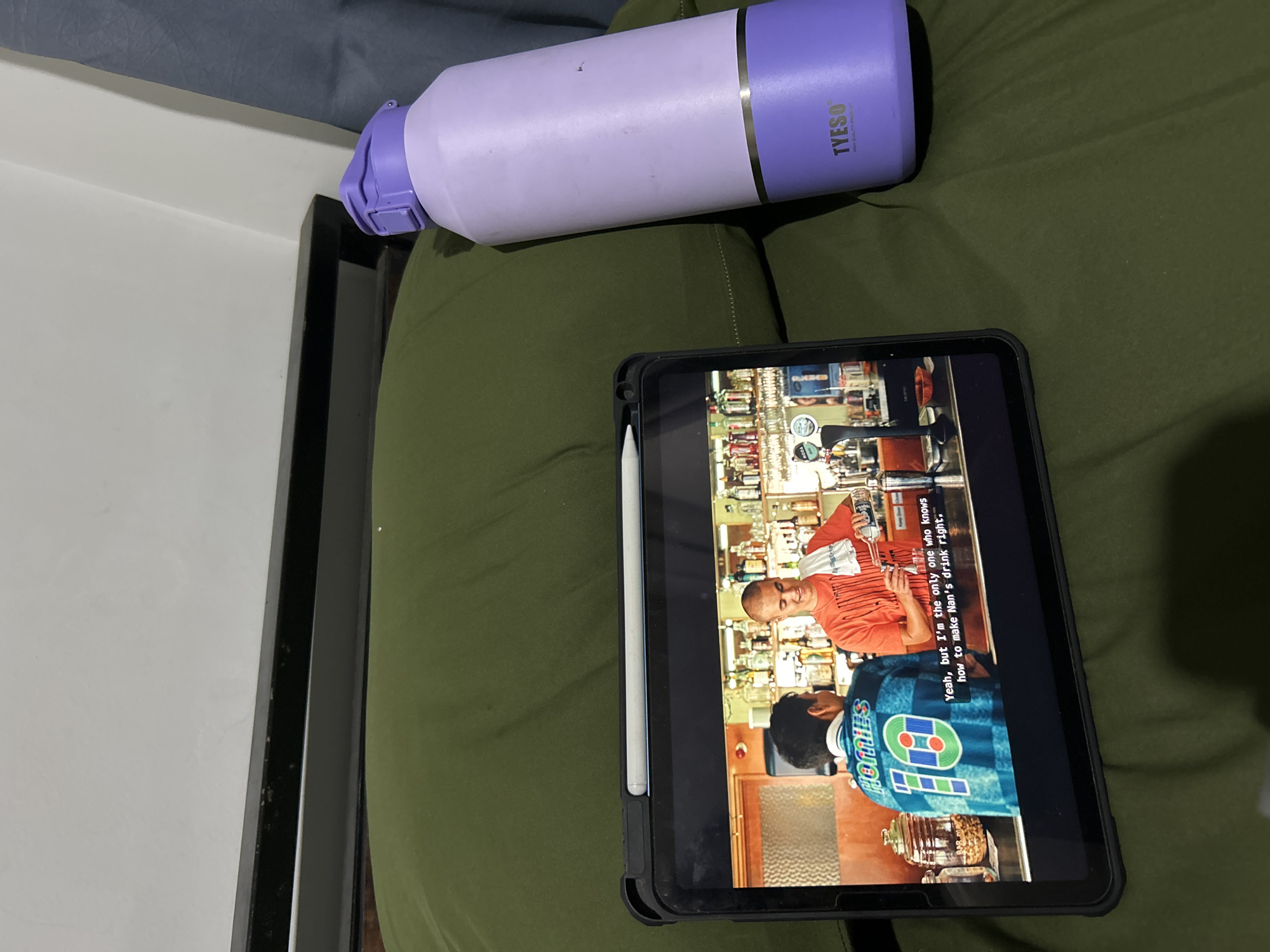 The image shows a close-up view of a tablet displaying a scene from a television show or movie. The screen depicts two individuals in what appears to be a bar or restaurant setting, with shelves stocked with bottles in the background. One person, wearing an orange shirt, is holding a bottle and smiling. The other person, in a blue checkered jacket, is seen from the back. Subtitles are visible at the bottom of the screen, reading "Yeah, but I'm the only one who knows how to make Nan's drink right."
Above and to the left of the tablet, a purple and light-purple water bottle with a silver band is placed on a green fabric surface. The water bottle has text on it that reads "TYESO HIGH QUALITY PRODUCT". The overall scene is dimly lit, suggesting it might be nighttime or an indoor setting. The green fabric appears to be a couch or bedding. The image seems to be taken in a home environment, possibly in Sukawati, Indonesia, as per the context provided.