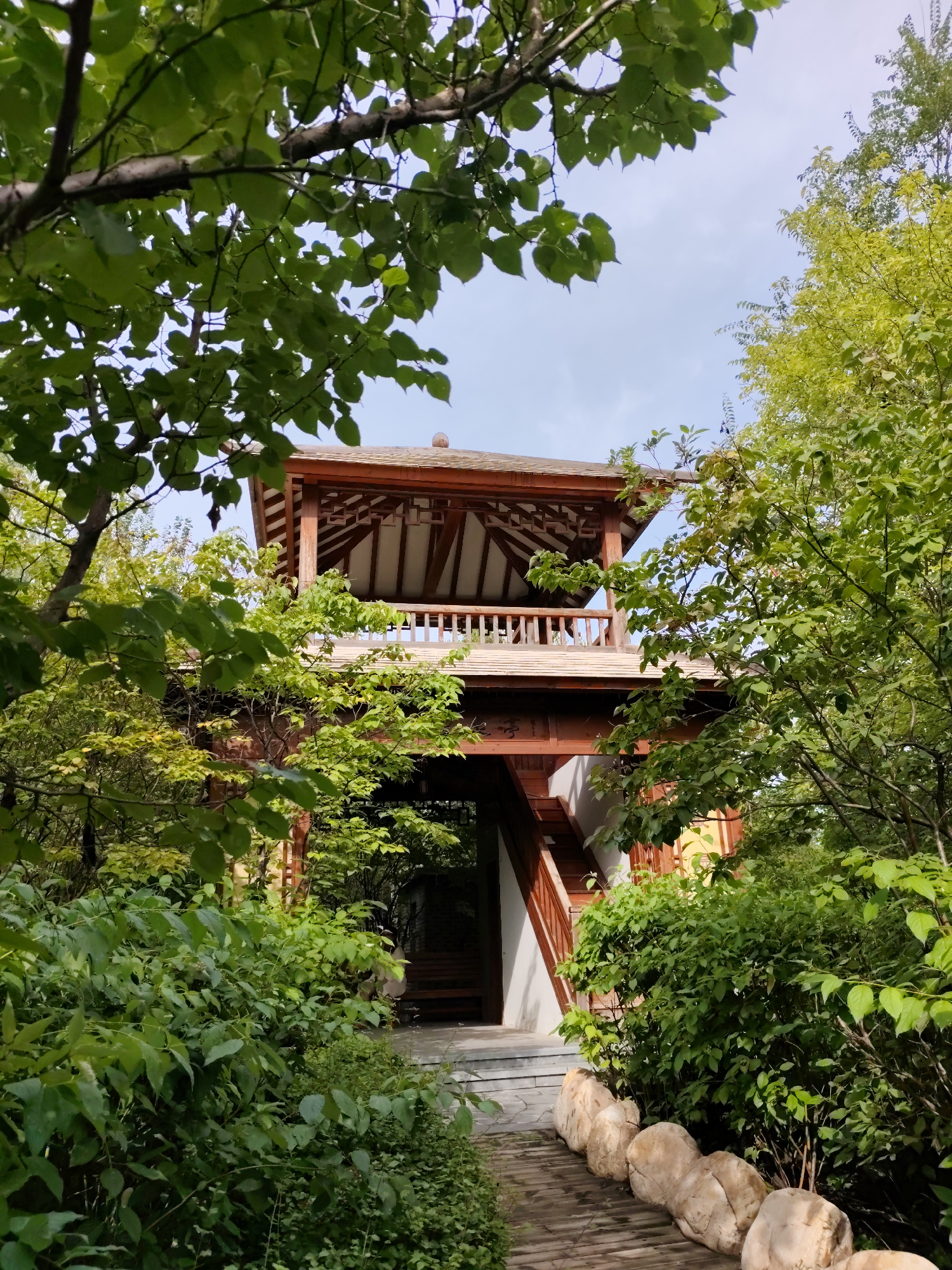 The image shows a traditional Chinese-style wooden tower, partially obscured by lush green foliage, nestled within a garden setting. The tower features multiple levels with exposed wooden beams and a tiled roof. A wooden walkway, bordered by large, rounded stones, leads towards the base of the tower. Stairs can be seen ascending the side of the structure, and a railing encircles the upper level. The sky above is a light blue with scattered clouds. The scene appears to be located in Lindian, China, given the architectural style and the natural environment. There are no people or animals visible in the image.