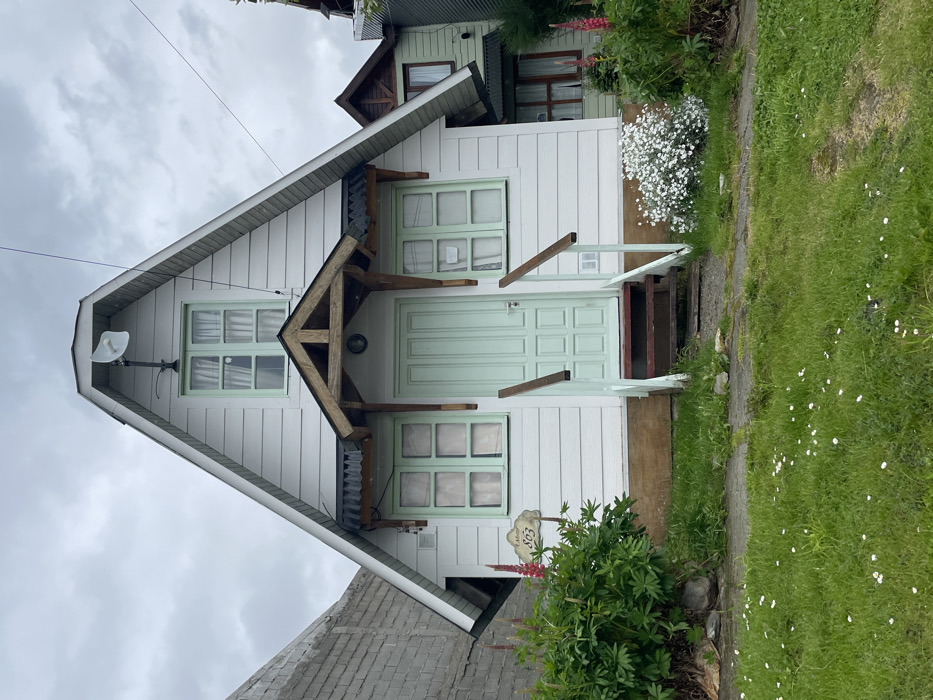 A-frame house with white siding and mint green trim, door, and windows. A wooden A-frame porch overhangs the front door. A small satellite dish is mounted on the left side of the house. A small sign with the number "803" is near the front. A patch of white flowers grows on the right side. Green grass with white daisies is visible in the foreground. The photo appears to be rotated, positioning the house sideways.